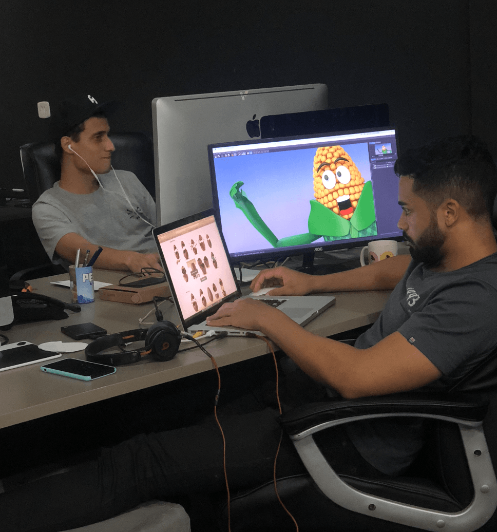 Dois homens trabalhando em mesas. Um edita em um laptop, enquanto um monitor maior exibe um personagem de desenho animado de milho.
