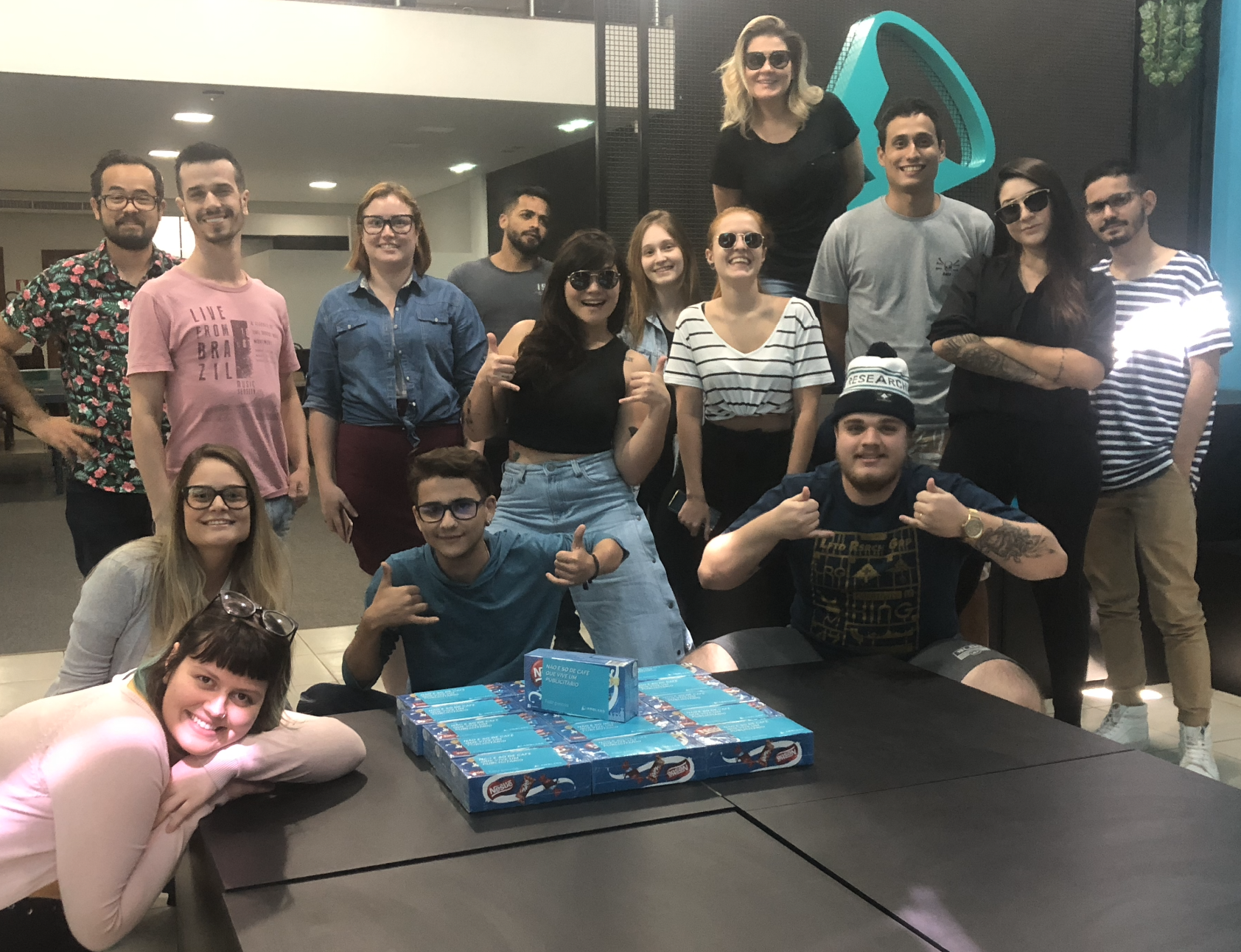 Um grupo de pessoas diversas sorrindo e posando ao redor de uma mesa com um presente. Elas estão em um ambiente de escritório, com um logotipo azul-petróleo visível ao fundo.
