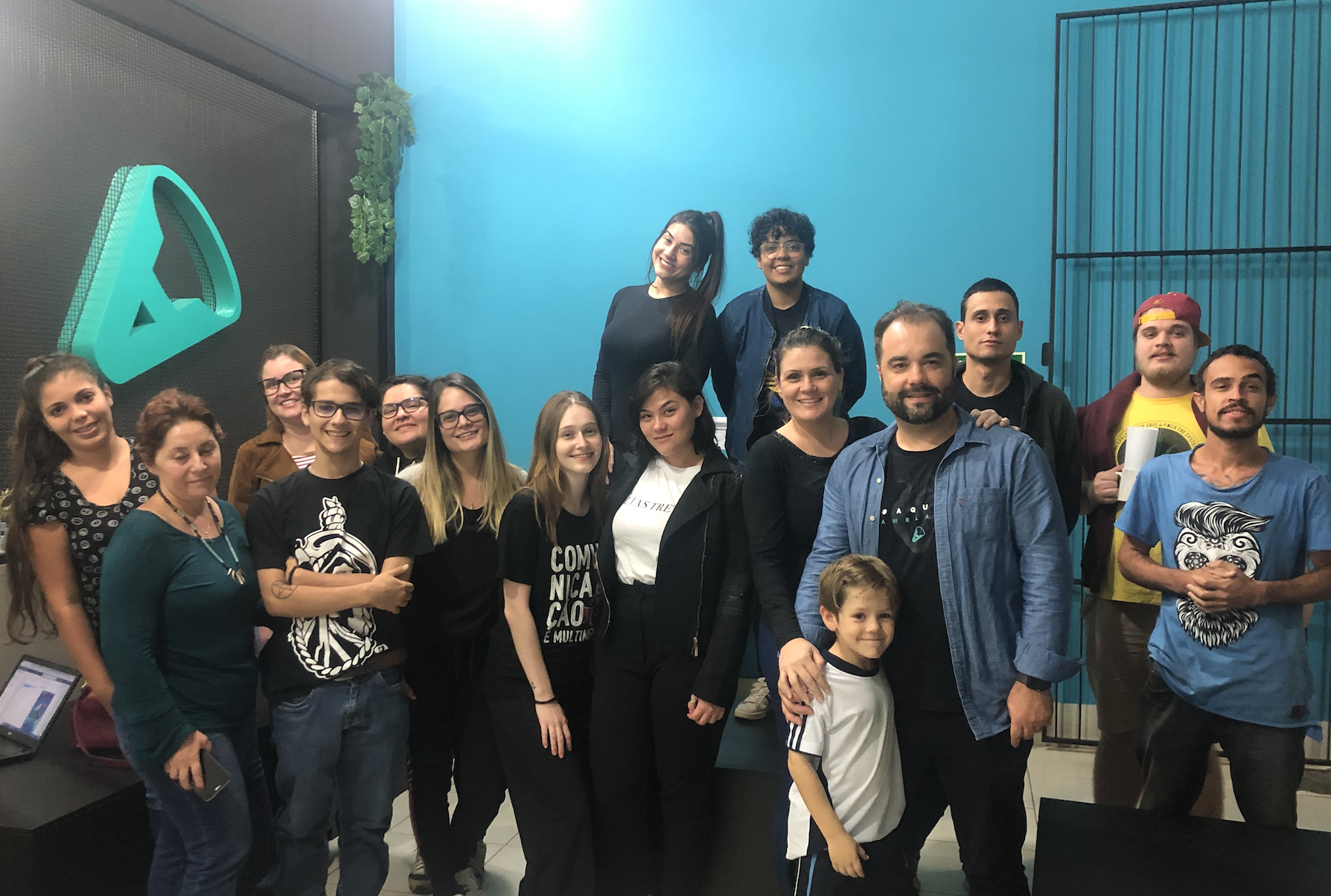 Grupo de pessoas sorrindo e posando juntas em frente a uma parede azul-petróleo, algumas segurando bebidas.