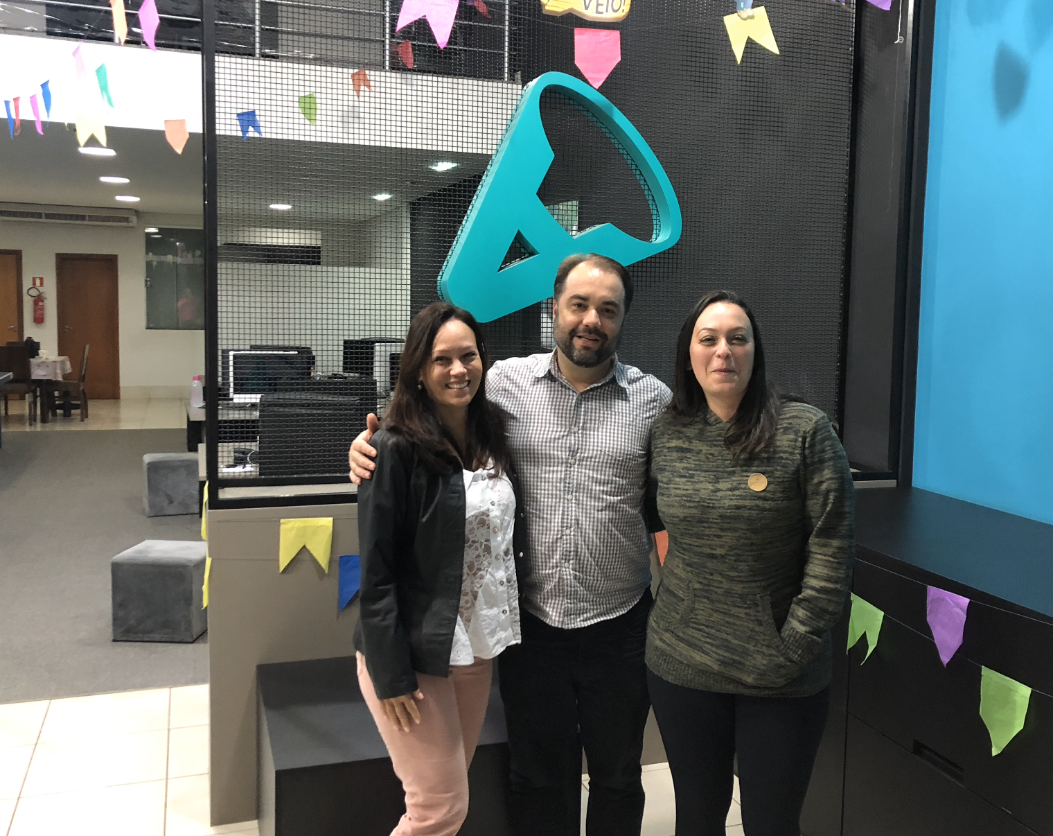 Três pessoas posam para uma foto em um escritório. Um homem barbudo está entre duas mulheres, sorrindo. Decorações de festa são visíveis.