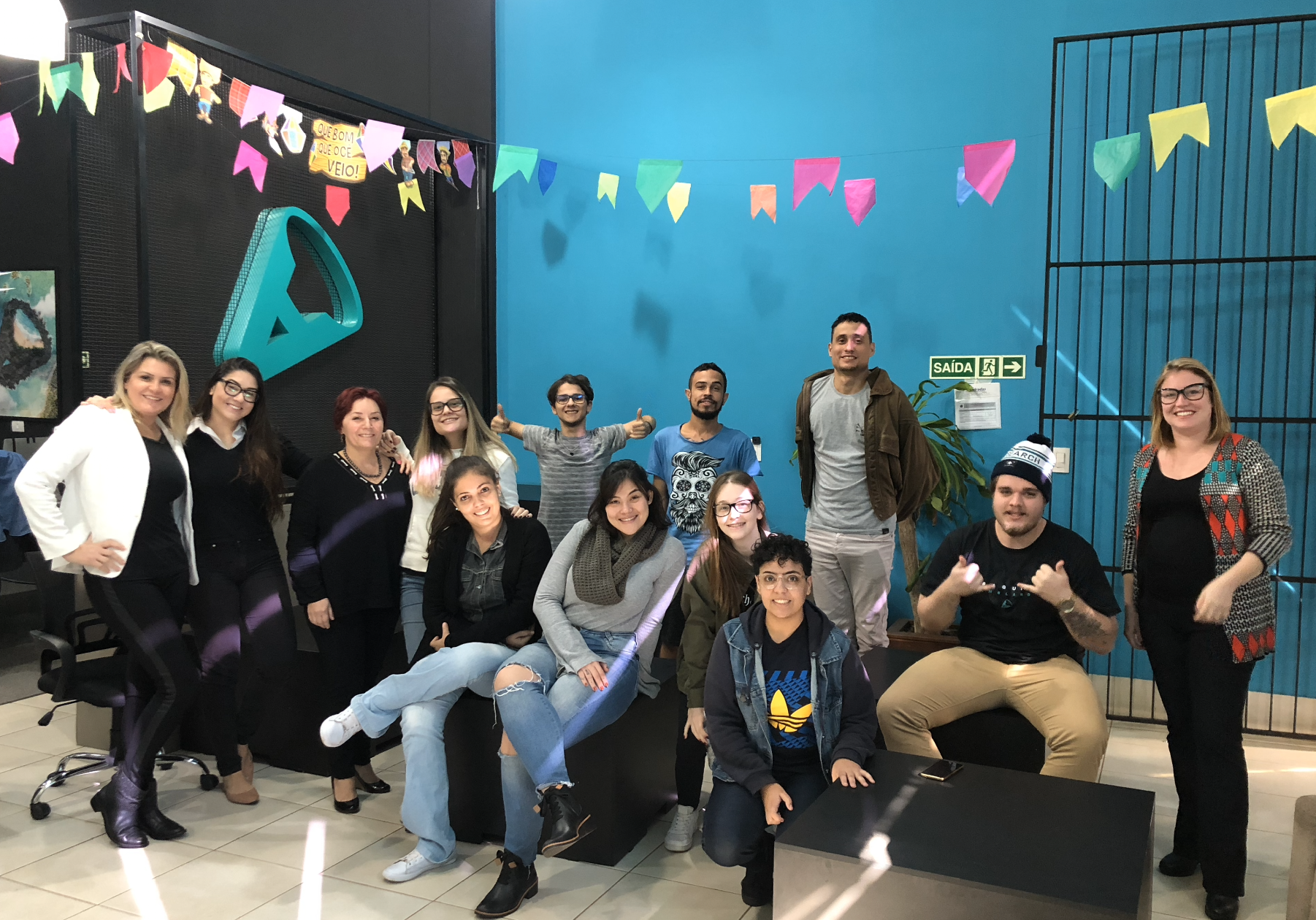 Grupo de pessoas posando para uma foto em um escritório bem decorado, algumas sorrindo, com uma parede azul ao fundo.