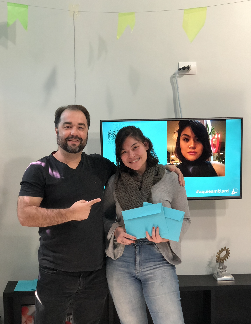 Um homem e uma mulher sorriem, um ao lado do outro. O homem aponta para a mulher, que segura uma caixa azul. Uma videochamada está sendo exibida em uma tela próxima.