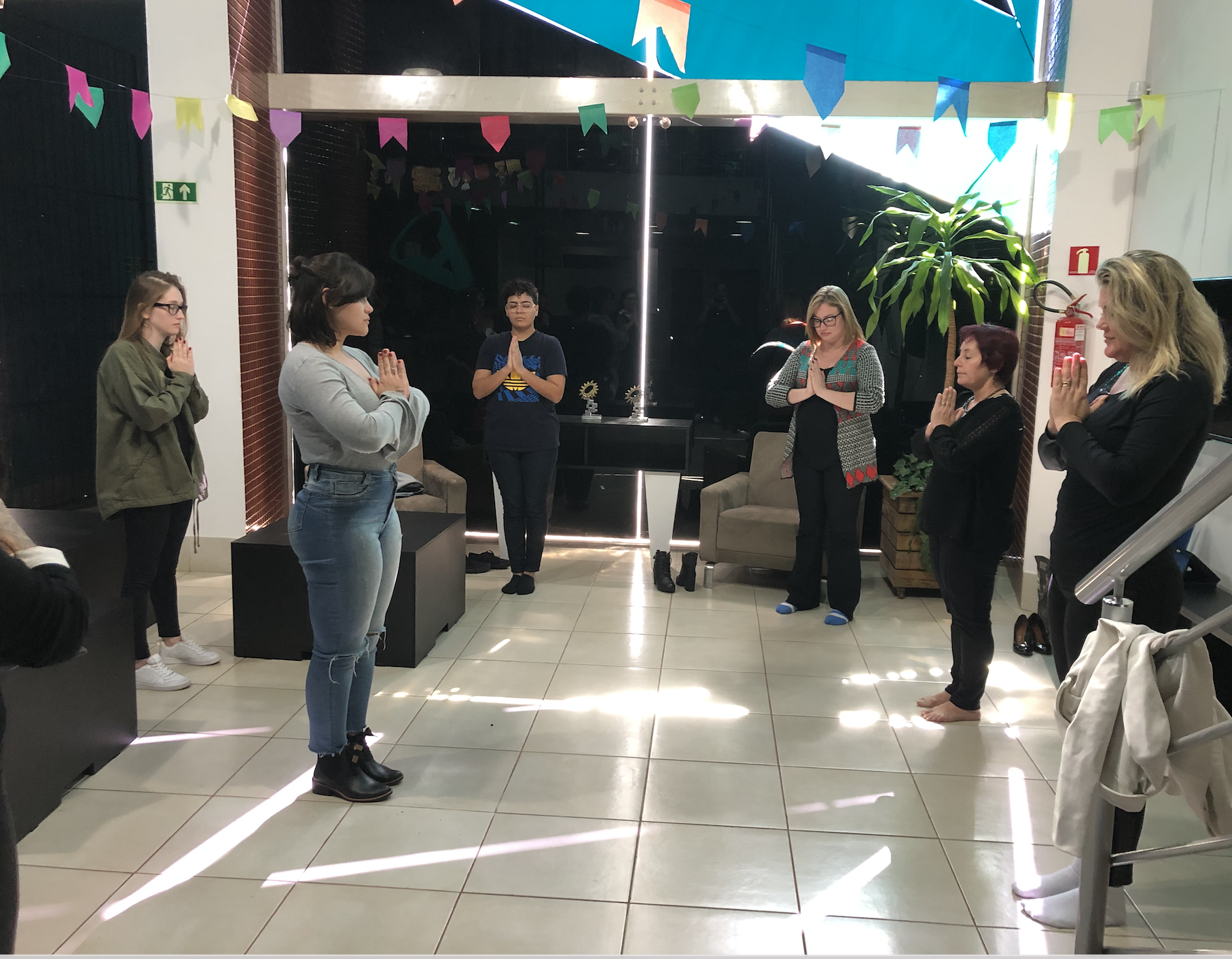 Grupo de pessoas em uma sala iluminada com as mãos juntas em oração, possivelmente uma sessão de meditação ou ioga.