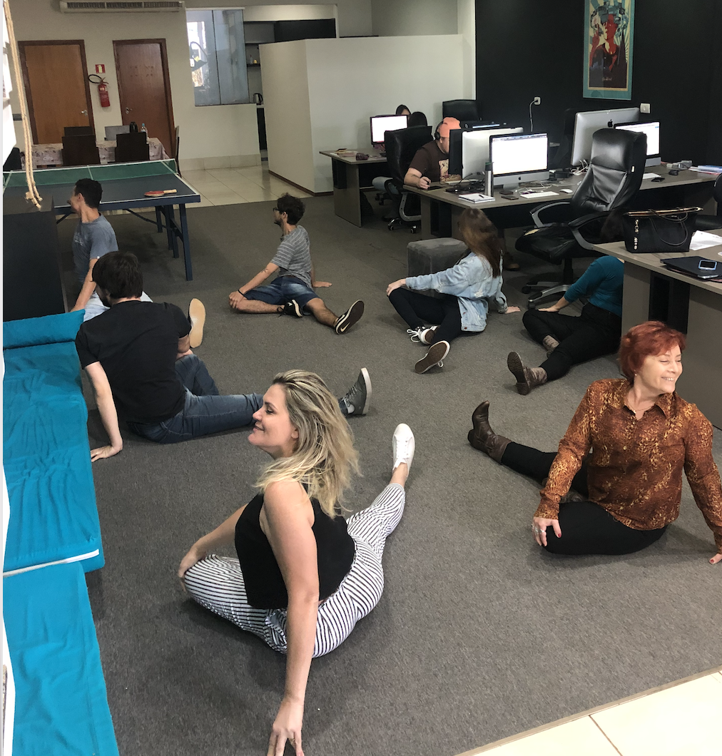 Pessoas se alongando no chão em um escritório. Uma mulher de calça estampada sorri enquanto faz um espacate. Outras pessoas se alongam atrás dela.