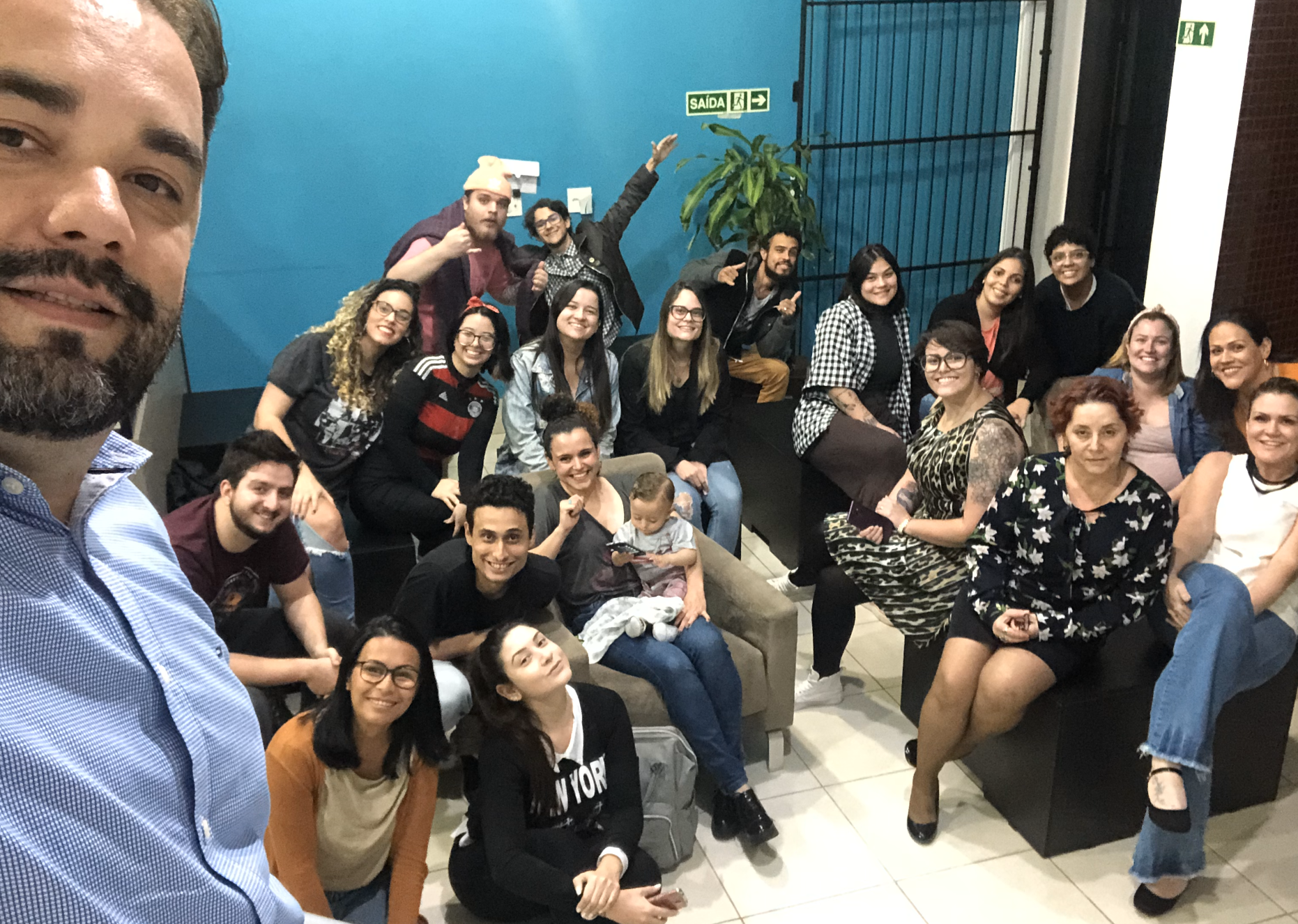 Foto de grupo de pessoas diversas sorrindo e posando em um ambiente interno, algumas sentadas em um sofá, com um homem tirando a selfie.