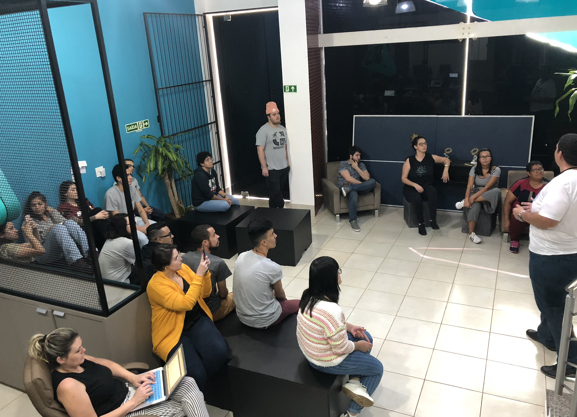 Um grupo de pessoas sentadas e em pé em um ambiente interno, algumas olhando para um homem que fala. A sala tem paredes azuis e brancas e móveis modernos.