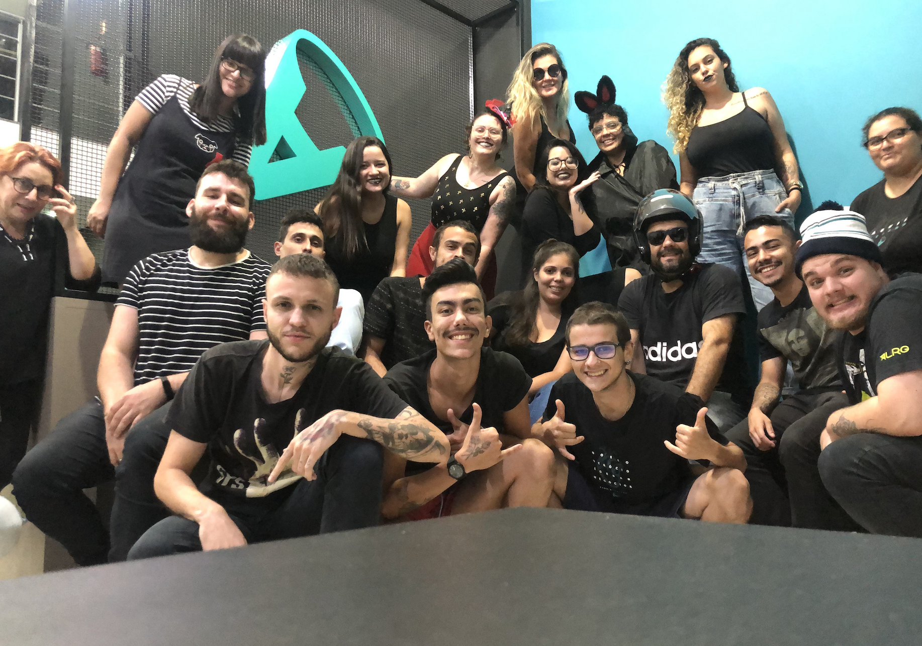 Grupo de pessoas posando em frente a uma parede azul-petróleo com um logotipo. A maioria sorri, algumas vestem roupas pretas.
