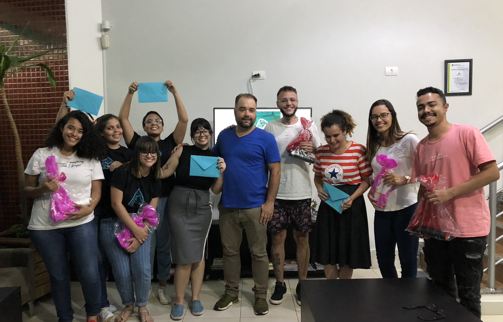 Grupo de pessoas sorrindo e segurando presentes, algumas segurando envelopes azuis. Provavelmente estão comemorando em um ambiente interno bem iluminado.