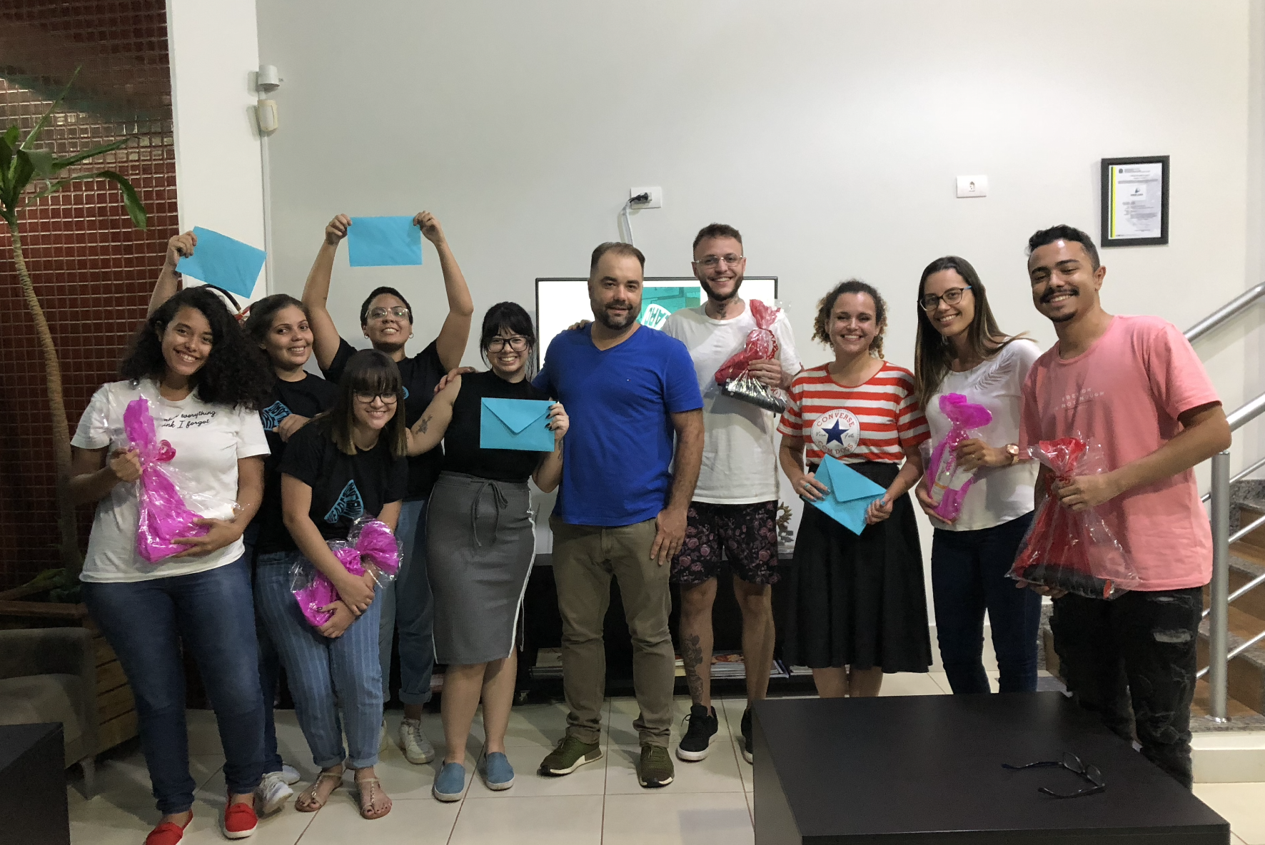 Grupo de pessoas sorrindo e segurando presentes e cartões azuis, reunidas em um ambiente interno bem iluminado.
