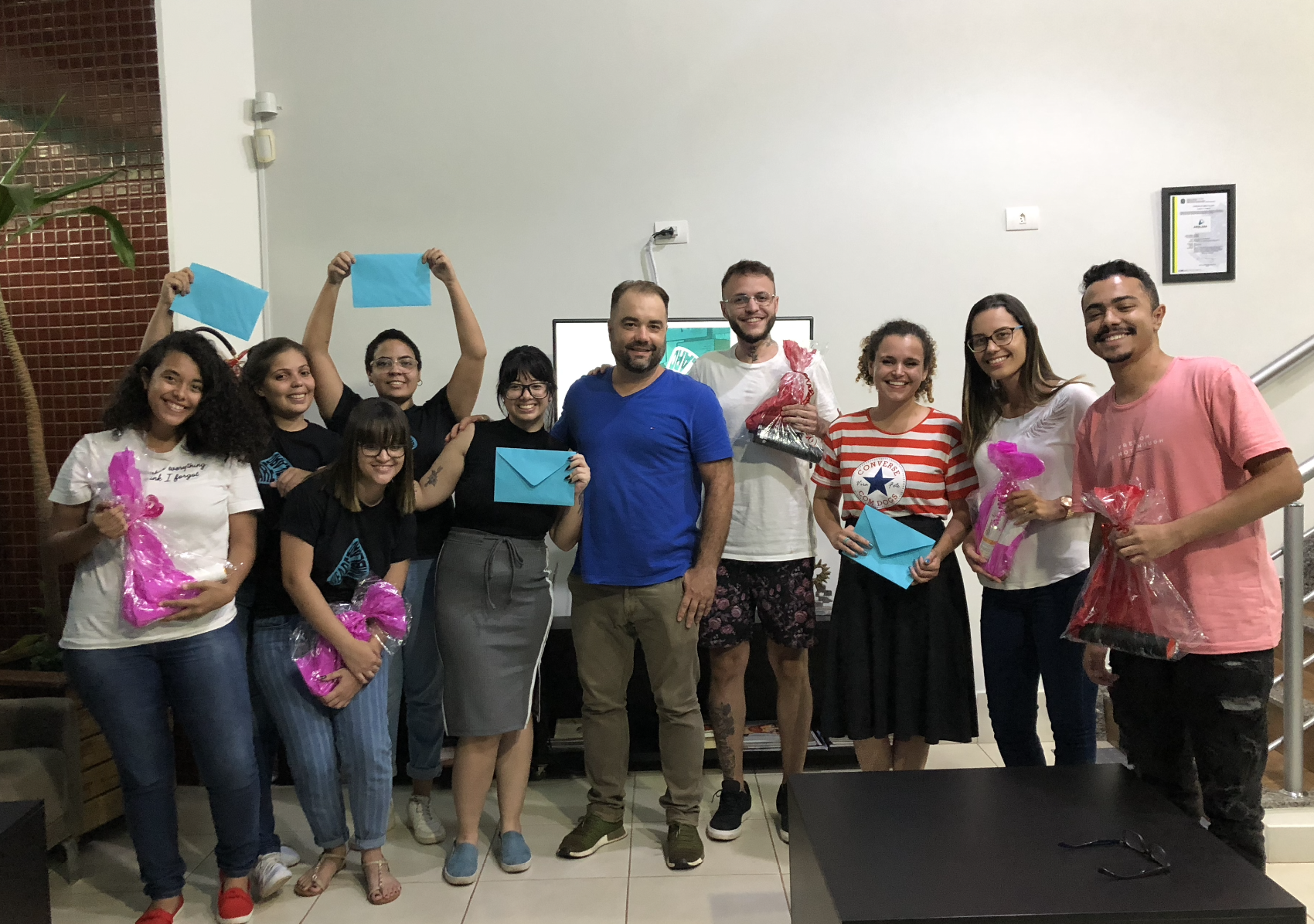 Um grupo de pessoas sorri, segurando presentes e envelopes azuis, em uma sala bem iluminada, provavelmente comemorando um evento.