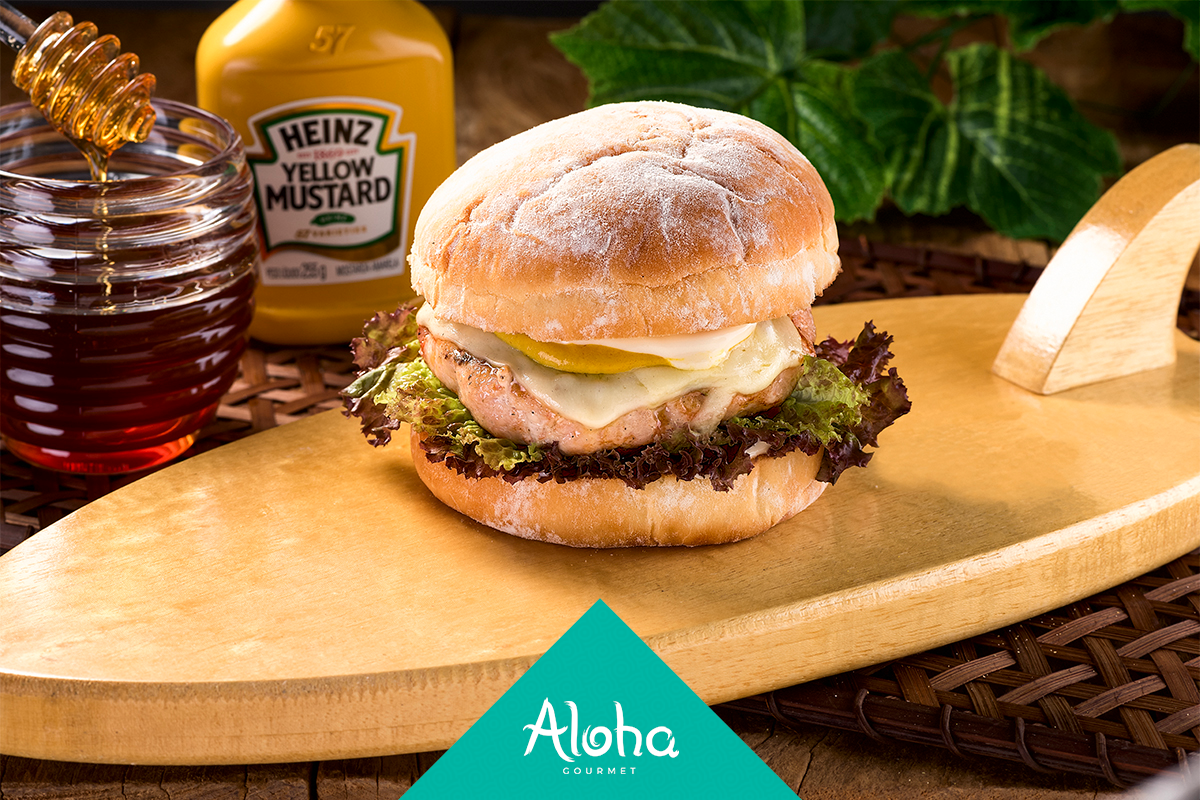 Hambúrguer em uma tábua de madeira em formato de prancha de surfe com mel e mostarda ao fundo; logotipo da Aloha na parte inferior.