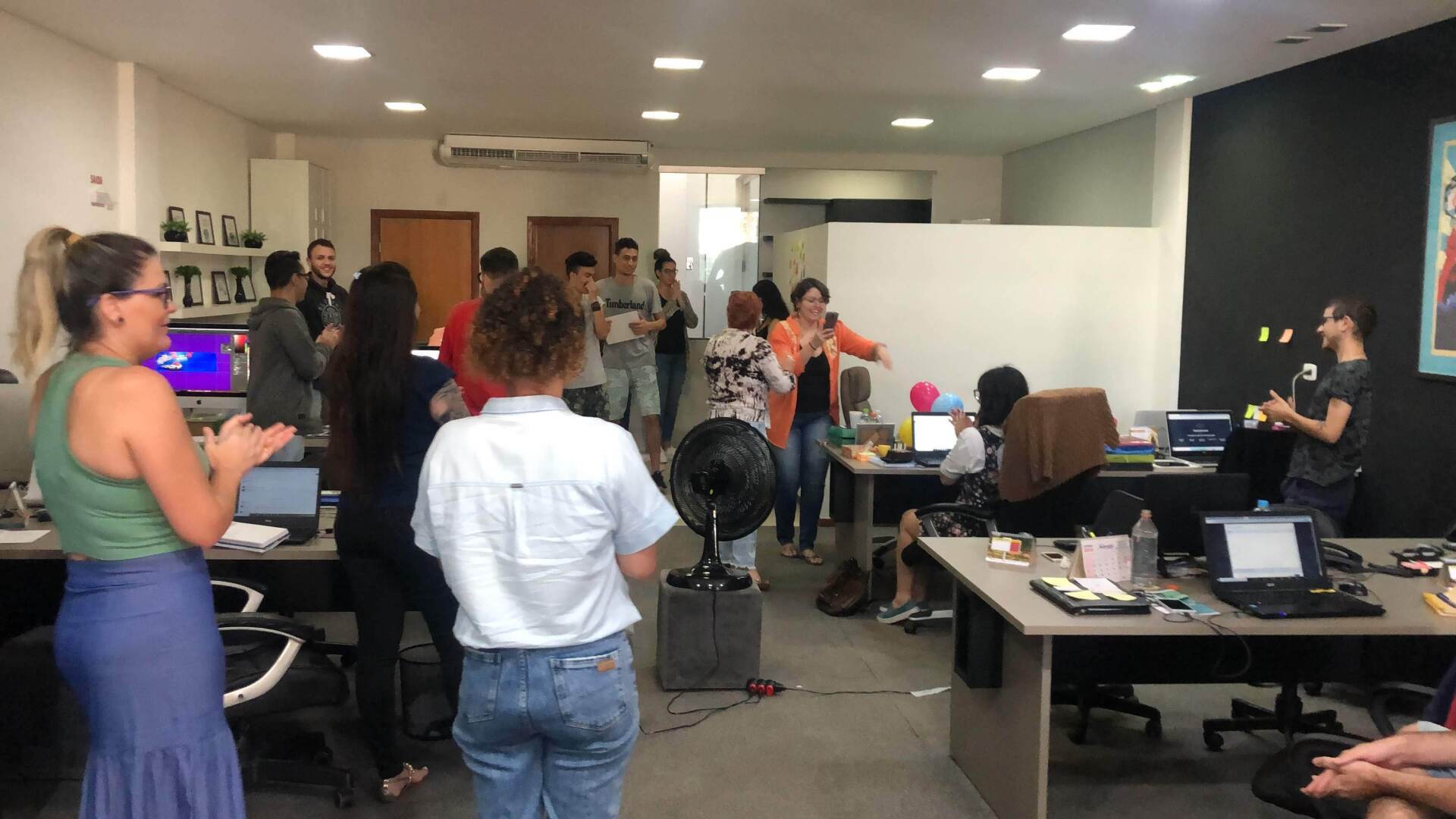 Pessoas comemorando em um escritório, batendo palmas e dançando. Um ventilador está sentado no centro da sala; laptops e mesas também são visíveis.