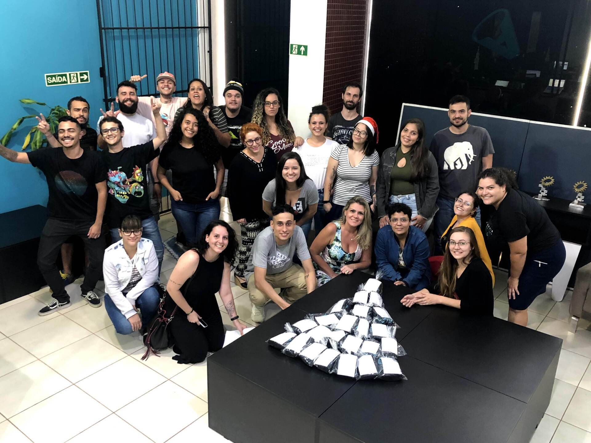 Foto de grupo com cerca de 30 pessoas ao redor de uma mesa preta com uma árvore de Natal de xícaras em um escritório. Pessoas sorrindo.