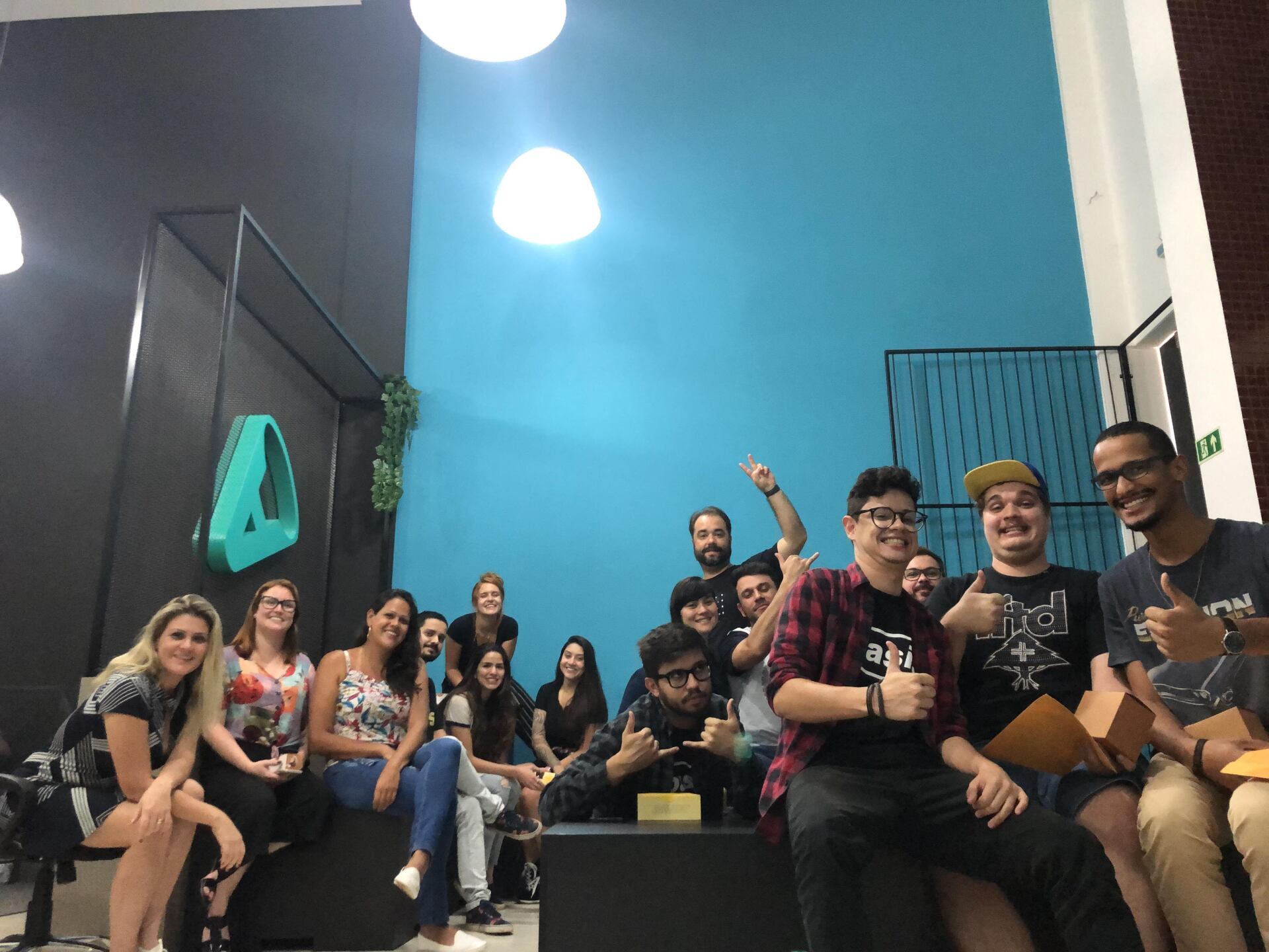 Grupo de pessoas sorrindo e comemorando juntas em um espaço moderno com paredes azuis e pretas, algumas fazendo sinal de positivo.