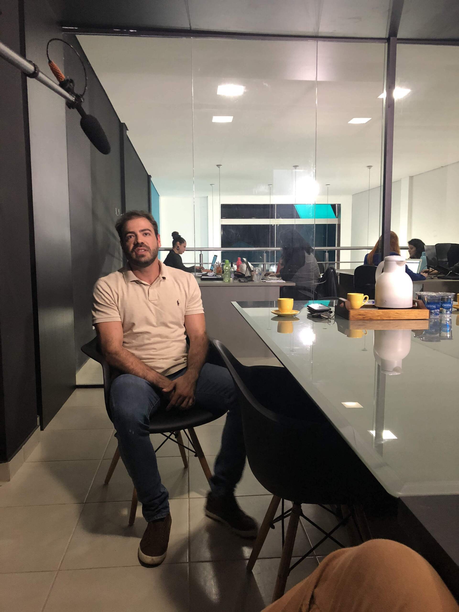 Um homem está sentado a uma mesa de conferências em um escritório, vestindo uma camisa bege e calça jeans. Outras pessoas estão ao fundo.