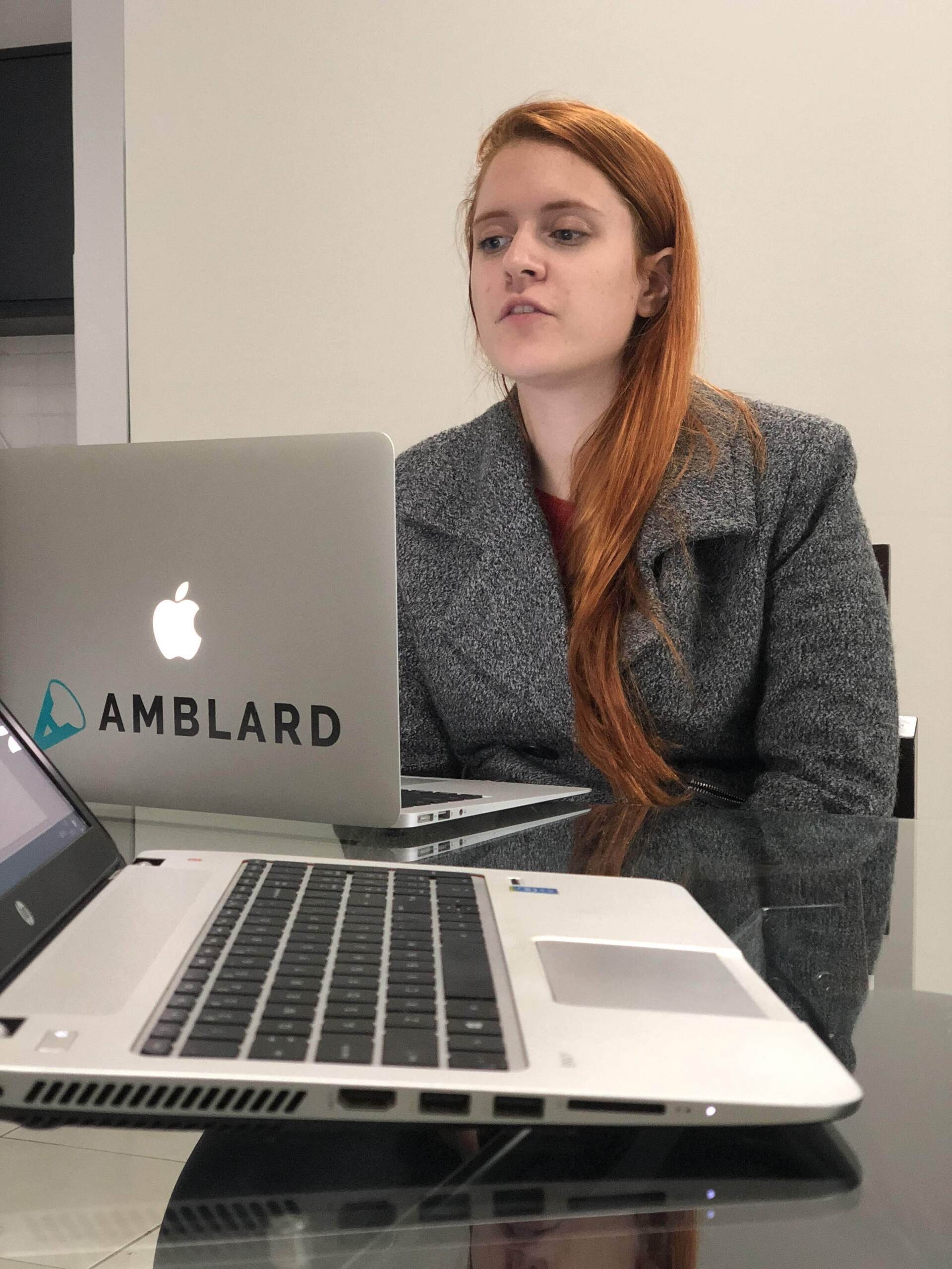 Uma mulher ruiva com um casaco cinza está sentada em frente a dois laptops em um escritório, olhando para uma tela.