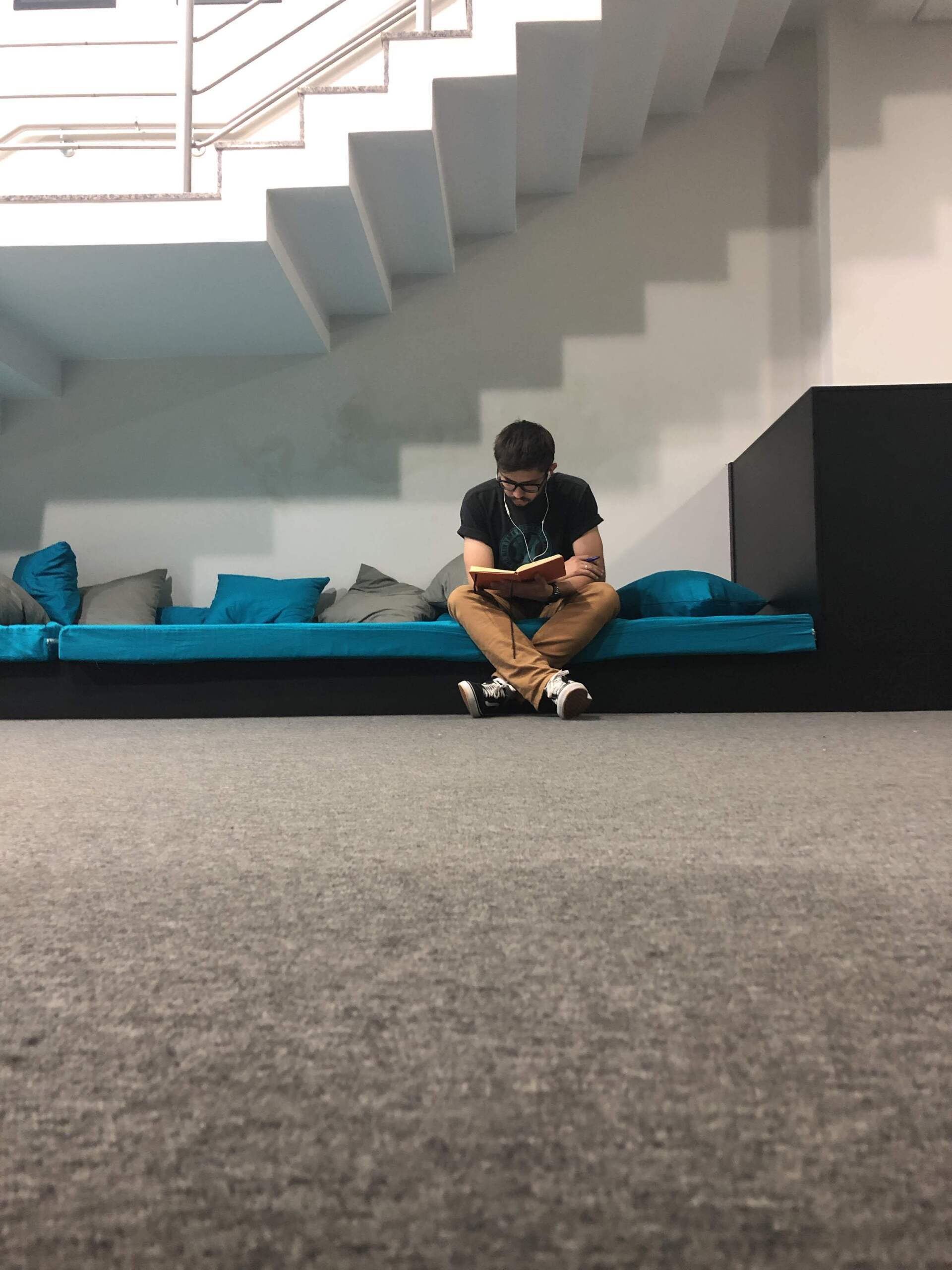 Homem lendo um livro sentado em uma área almofadada azul, embaixo de uma escada, em um prédio.