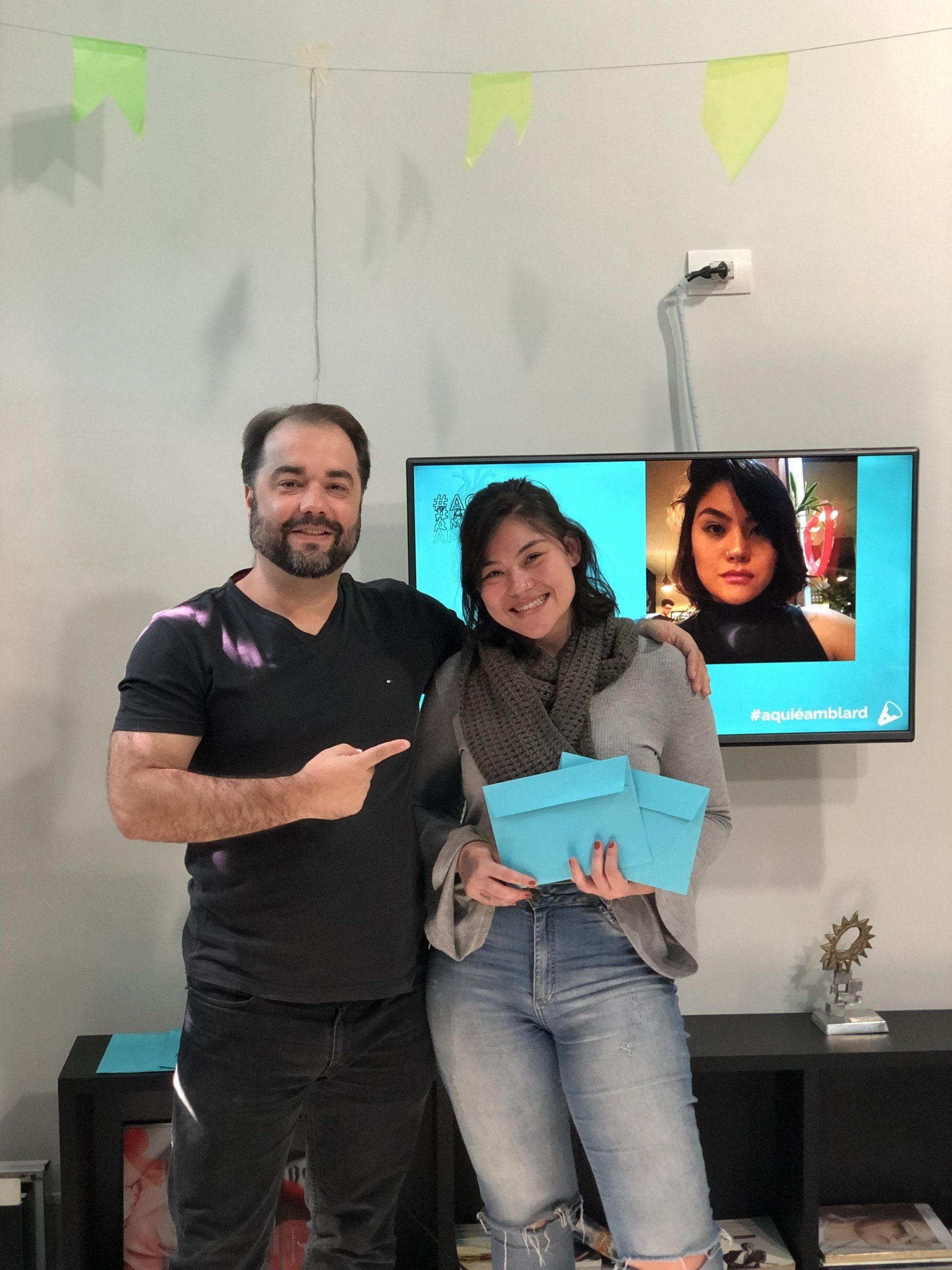 Um homem aponta para uma mulher sorridente segurando uma caixa azul. Eles estão perto de uma televisão exibindo a foto de uma mulher. Ao fundo, há um banner e uma prateleira.