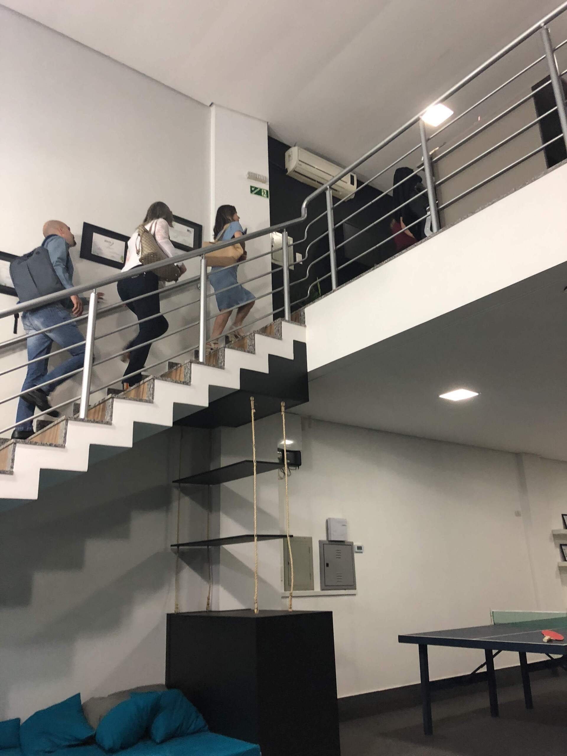 Pessoas subindo uma escada branca com corrimão de metal dentro de um escritório moderno. Um cubo preto e uma mesa de pingue-pongue também são visíveis.