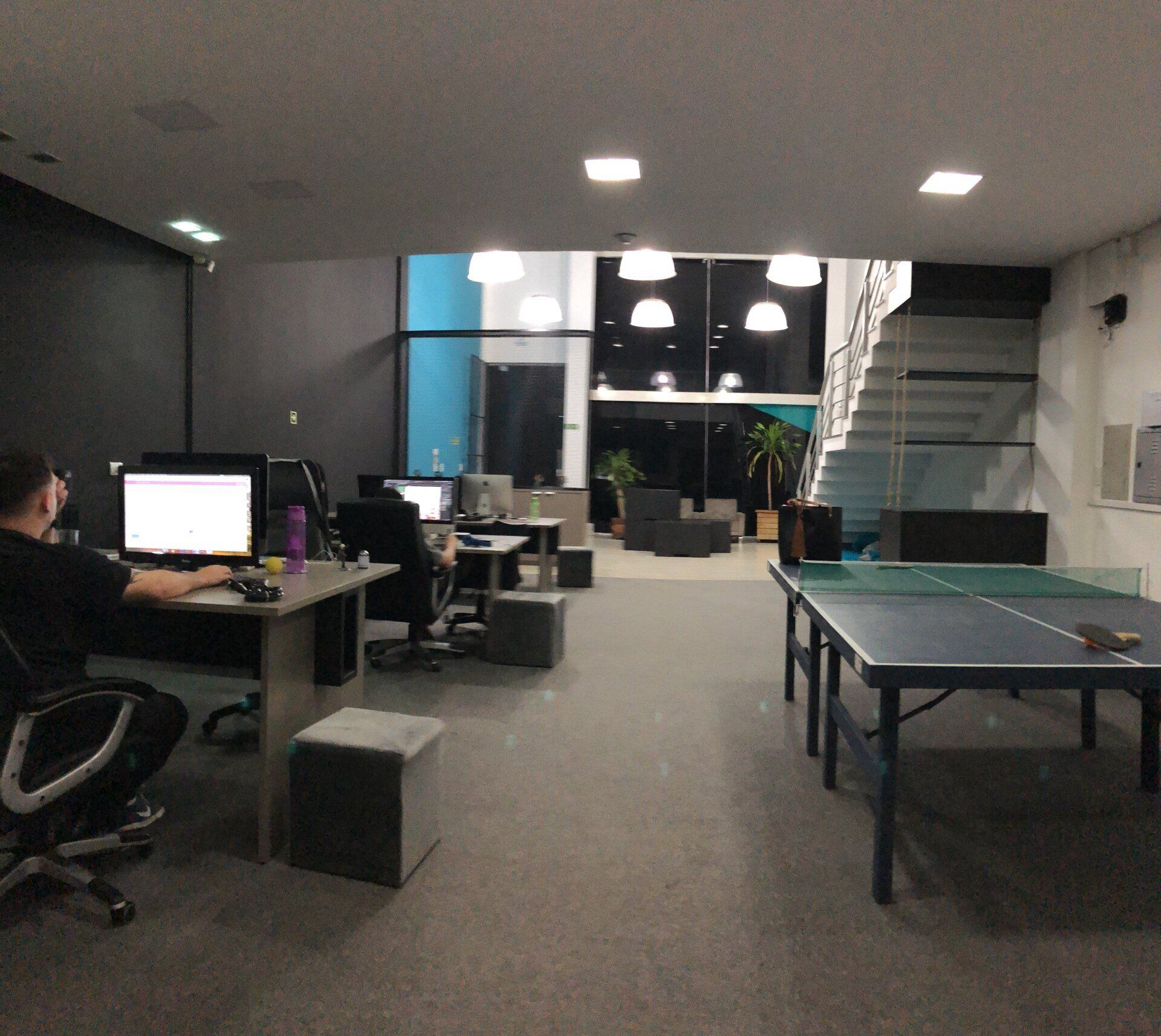 Interior de um escritório com pessoas trabalhando em computadores, uma mesa de pingue-pongue e uma escada. O espaço tem paredes cinza-escuras e iluminação forte no teto.