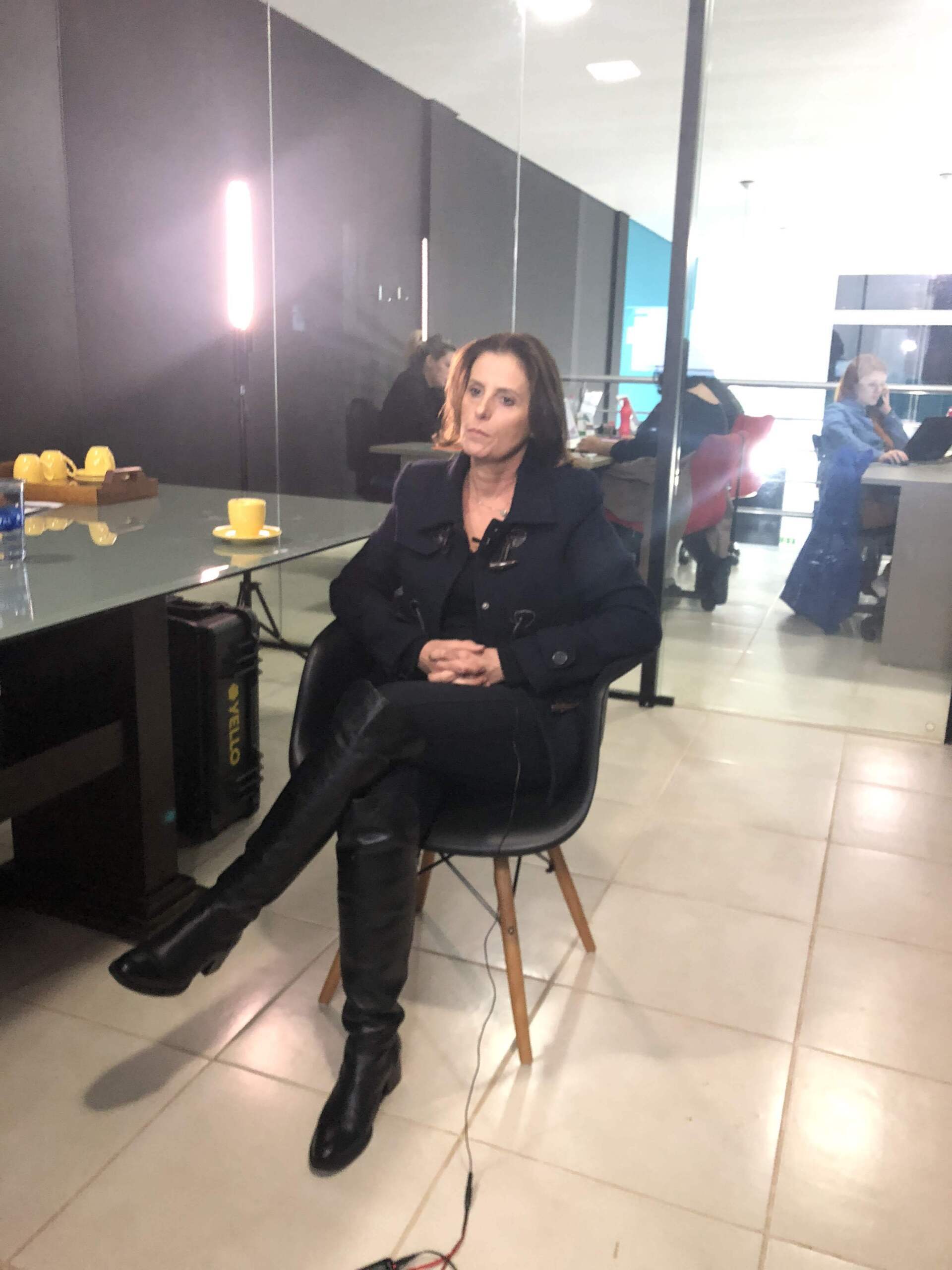 Mulher de casaco preto e botas sentada em uma cadeira em um ambiente interno. Ela tem as pernas cruzadas e uma expressão neutra. Iluminação e equipamentos ao fundo.