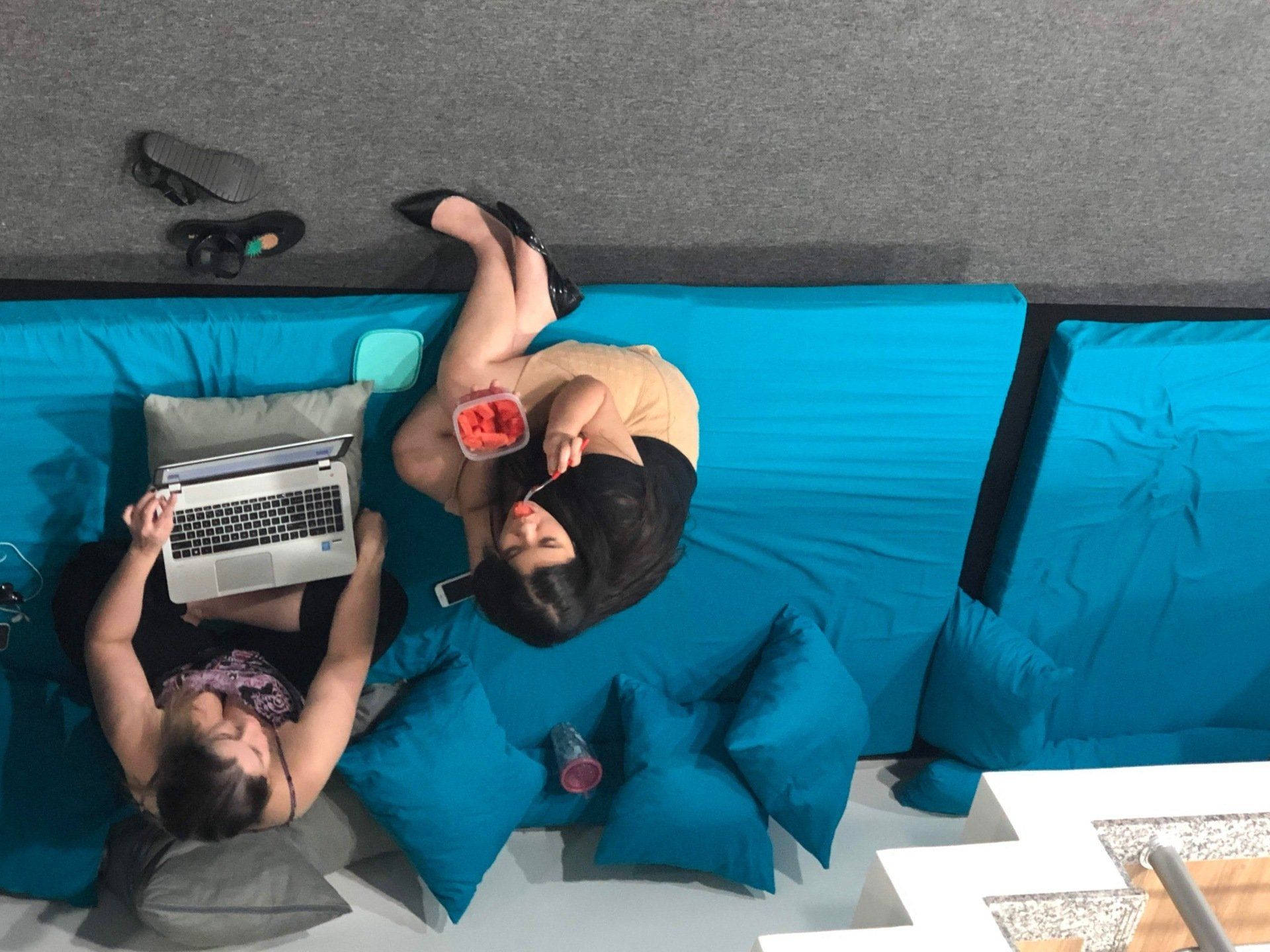 Duas mulheres em um sofá secional turquesa; uma trabalha em um laptop, a outra come morangos.