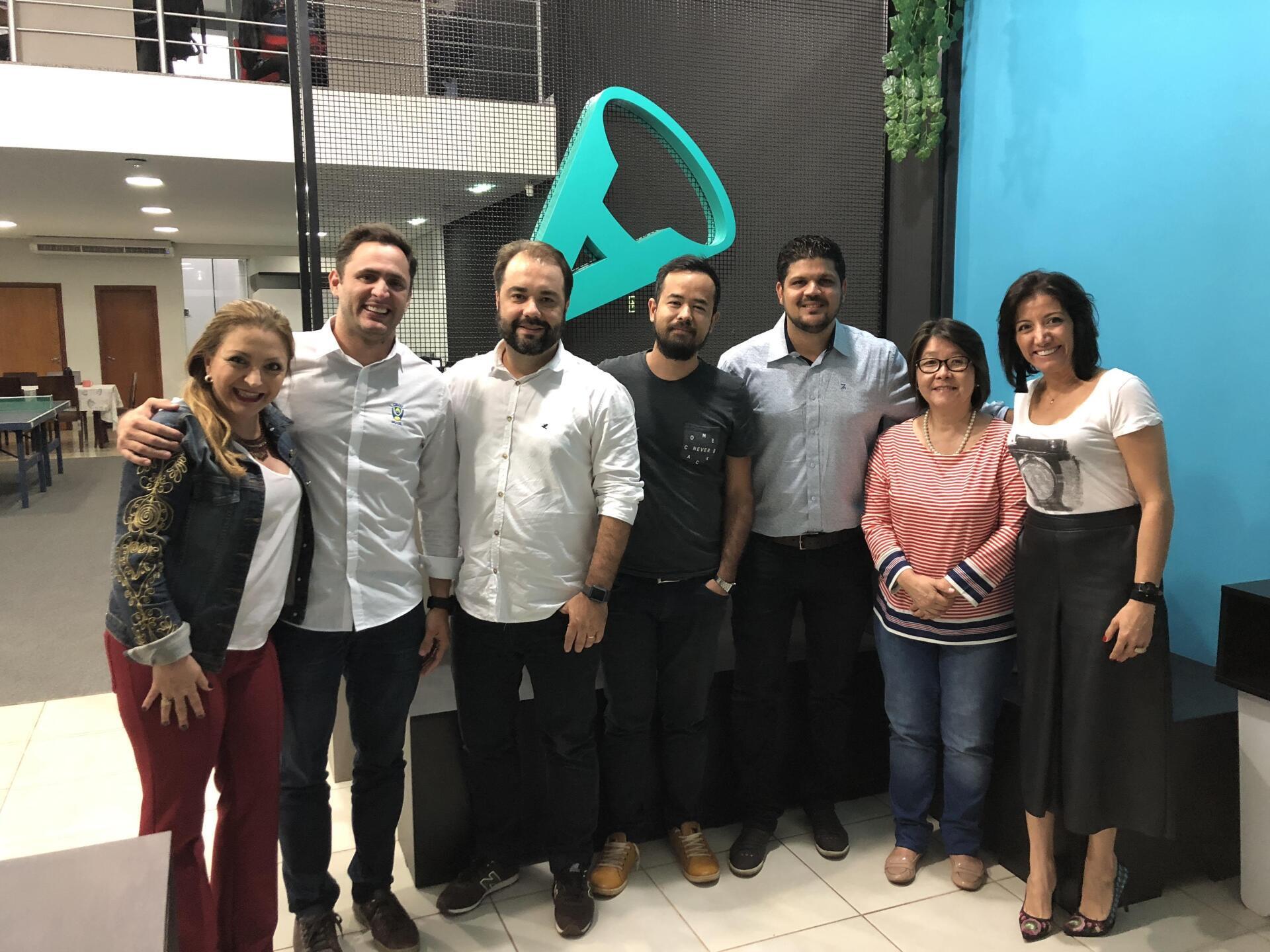 Um grupo de sete pessoas sorrindo e posando em um escritório com um logotipo na parede.