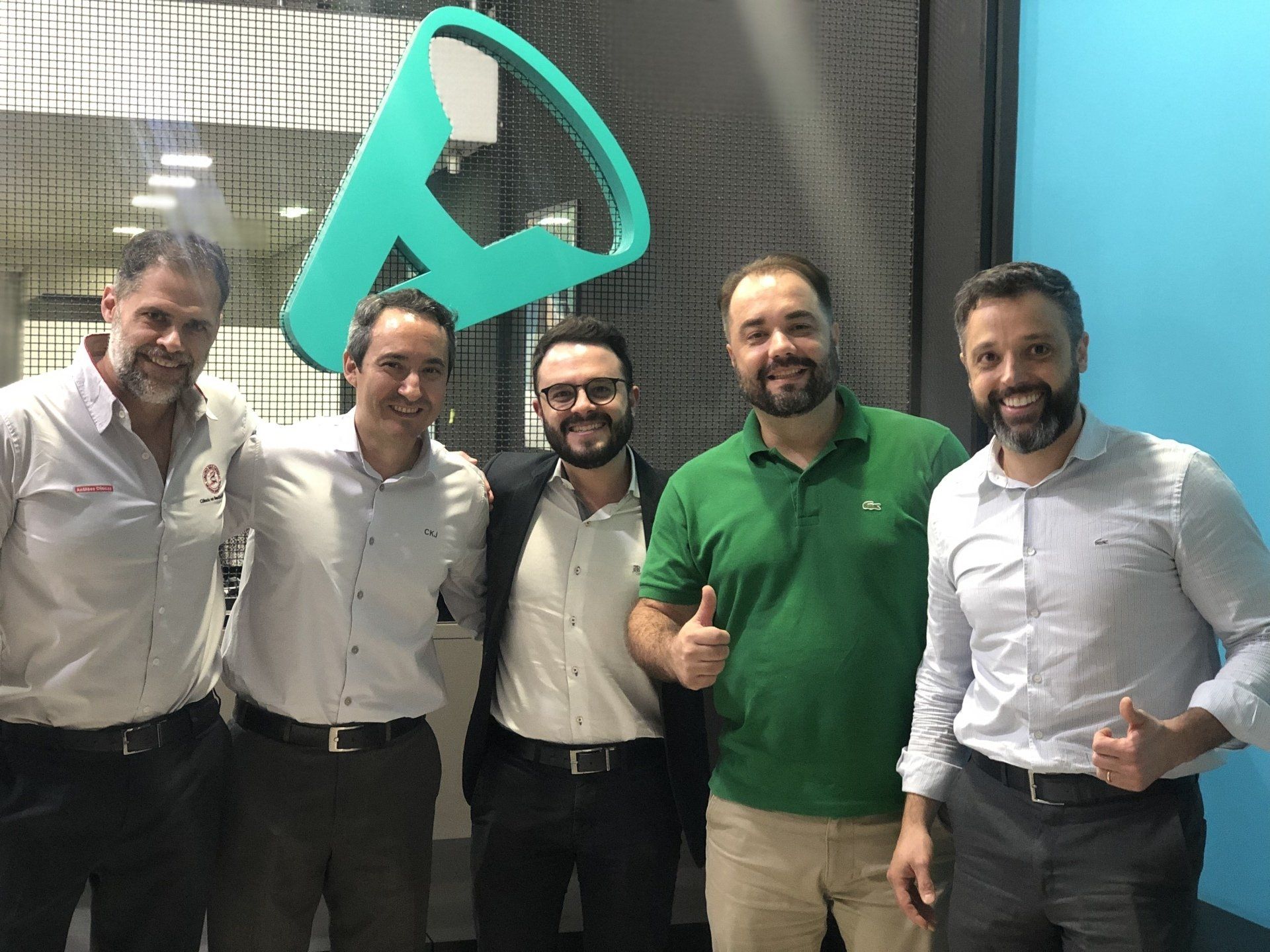 Cinco homens posando em um escritório com um logotipo na parede. Eles sorriem, alguns fazendo sinal de positivo.