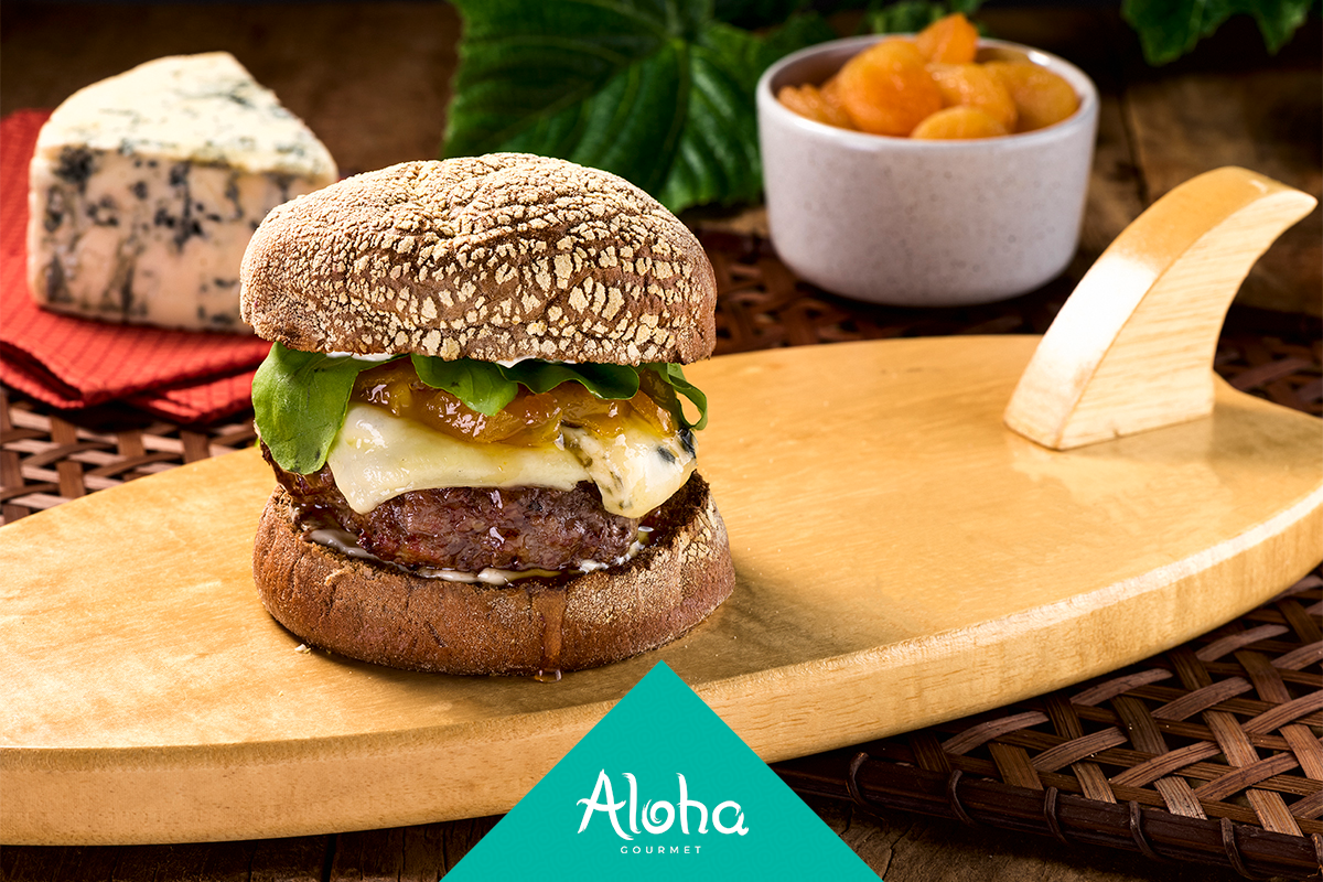 Hambúrguer com acompanhamentos em uma bandeja de madeira em formato de prancha de surfe, com queijo azul e frutas ao fundo.