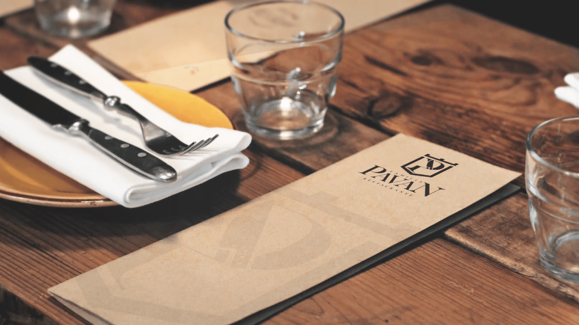 Mesa de madeira com cardápio de restaurante, talheres sobre guardanapo dobrado e copos. O cardápio tem a inscrição