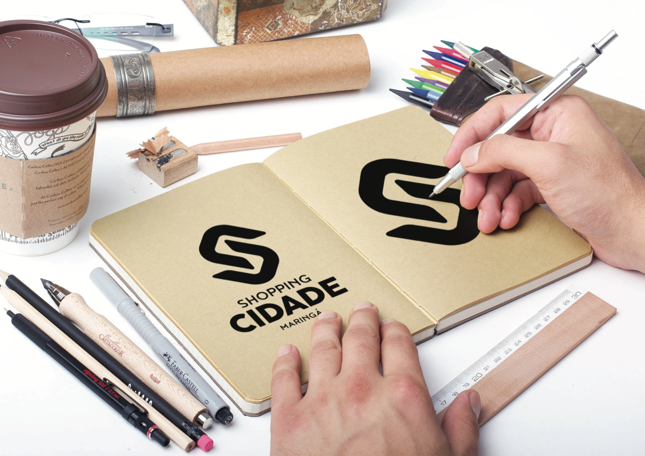 Esboço manual de um logotipo em um caderno. Xícara de café, lápis e régua sobre uma superfície branca. O logotipo diz