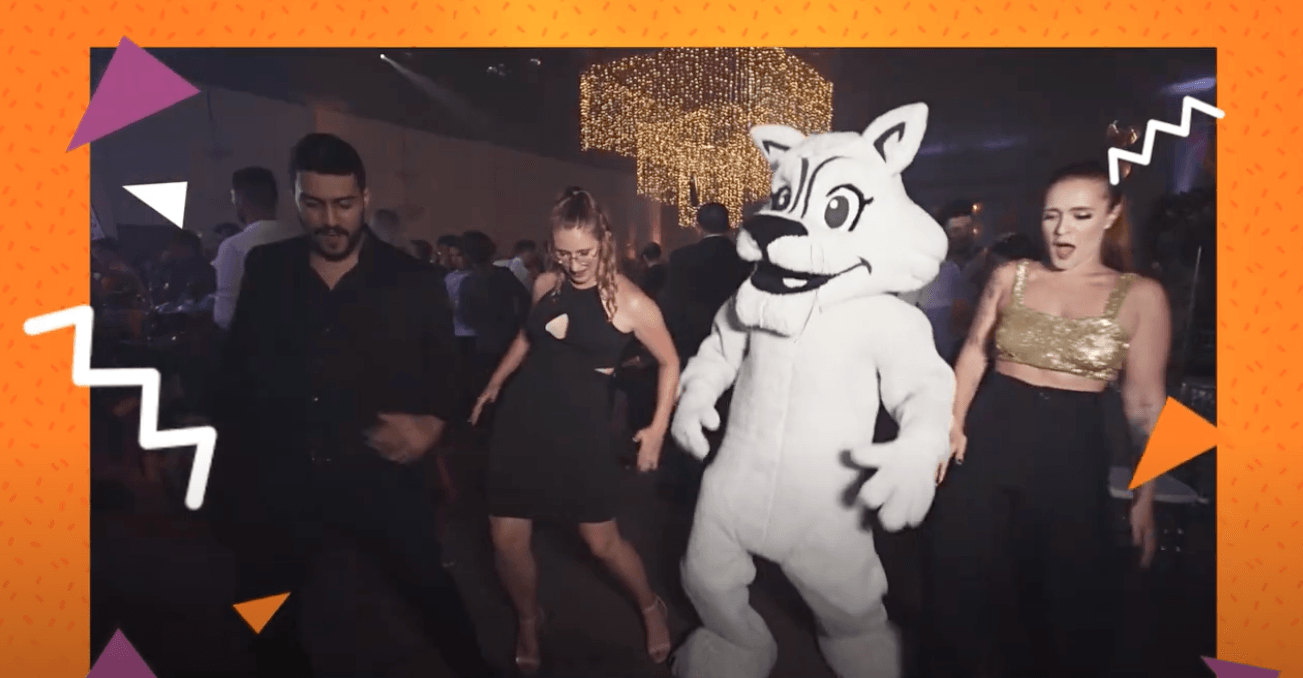 Pessoas dançando em uma festa com um mascote fantasiado de lobo branco; outros foliões ao fundo.