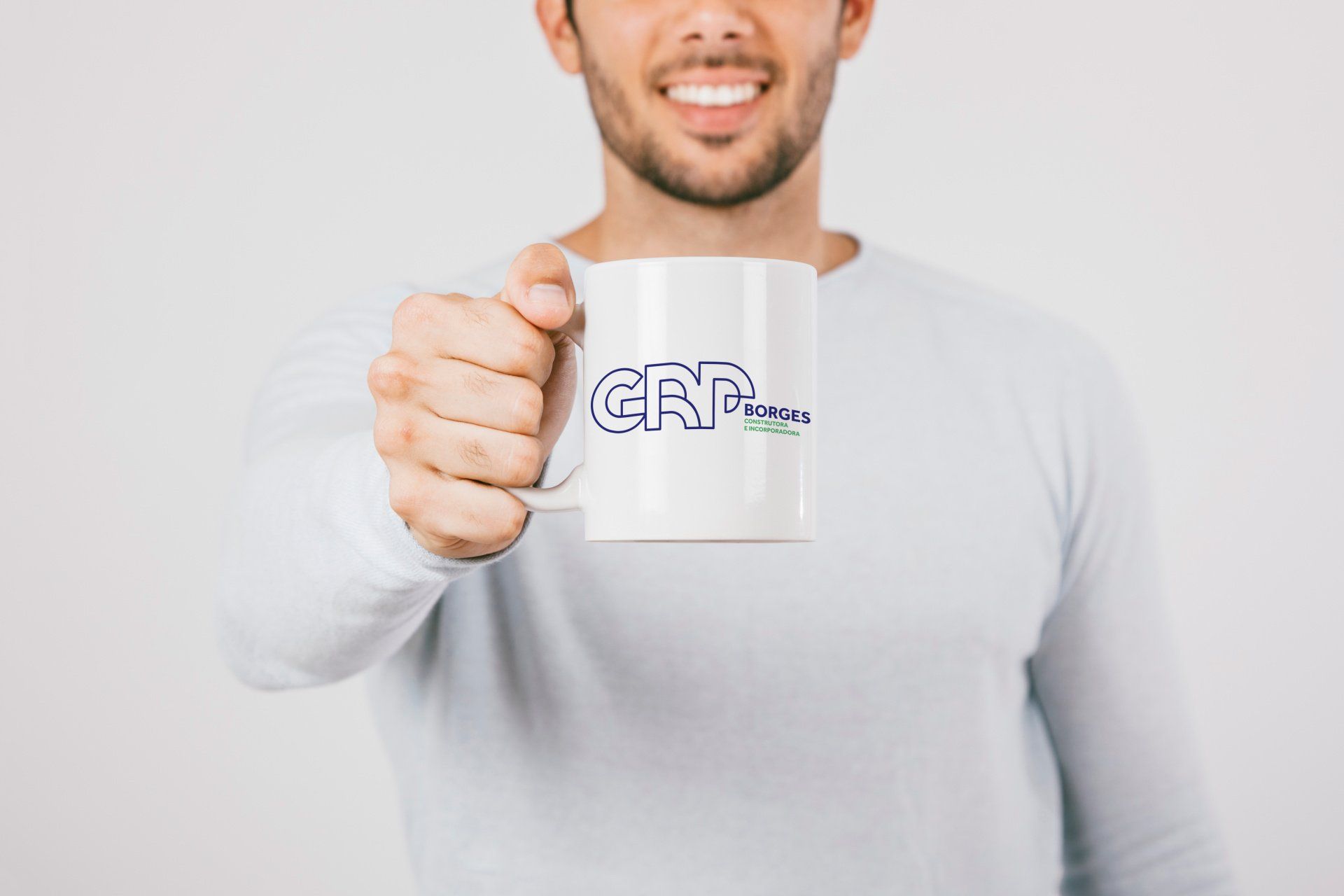 Homem sorrindo, segurando uma caneca branca com texto em azul e verde, provavelmente um logotipo, sobre um fundo claro.