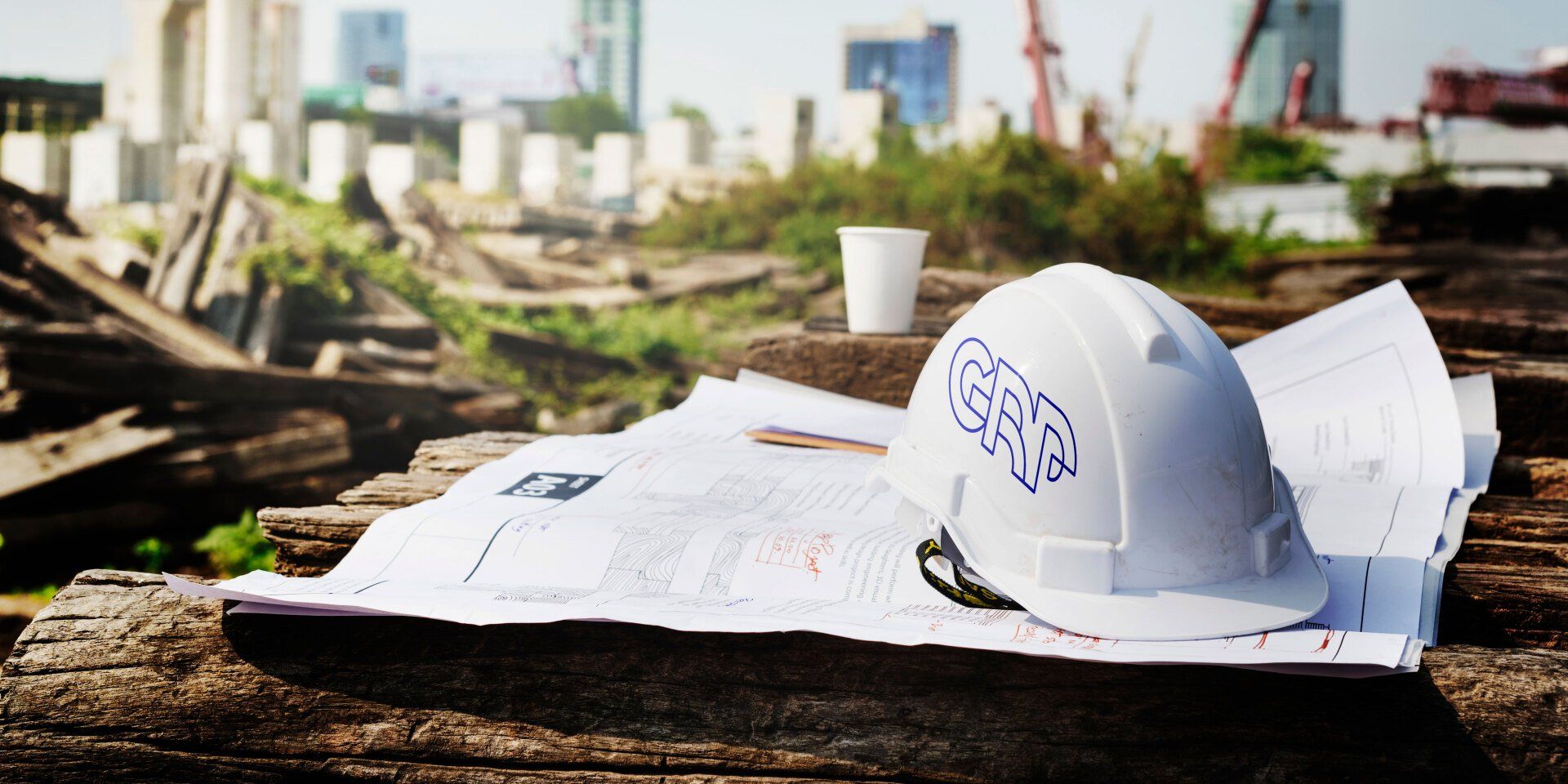 Canteiro de obras com um capacete branco, plantas e uma xícara de café em madeira desgastada, com edifícios ao fundo.