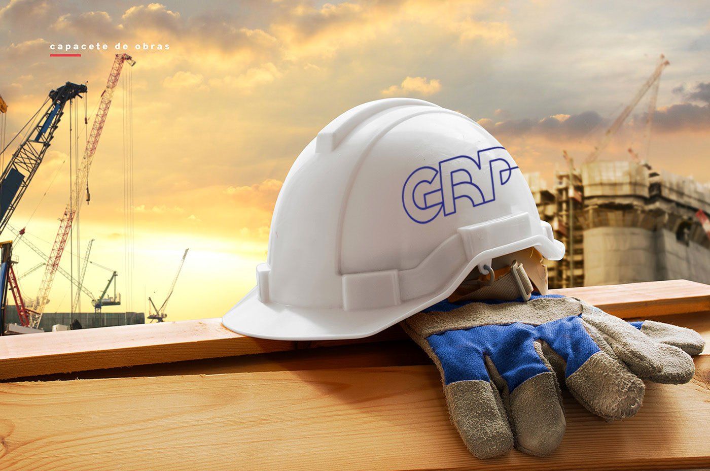 Capacete e luvas de trabalho sobre uma superfície de madeira, com guindastes de construção e um prédio em construção ao fundo.