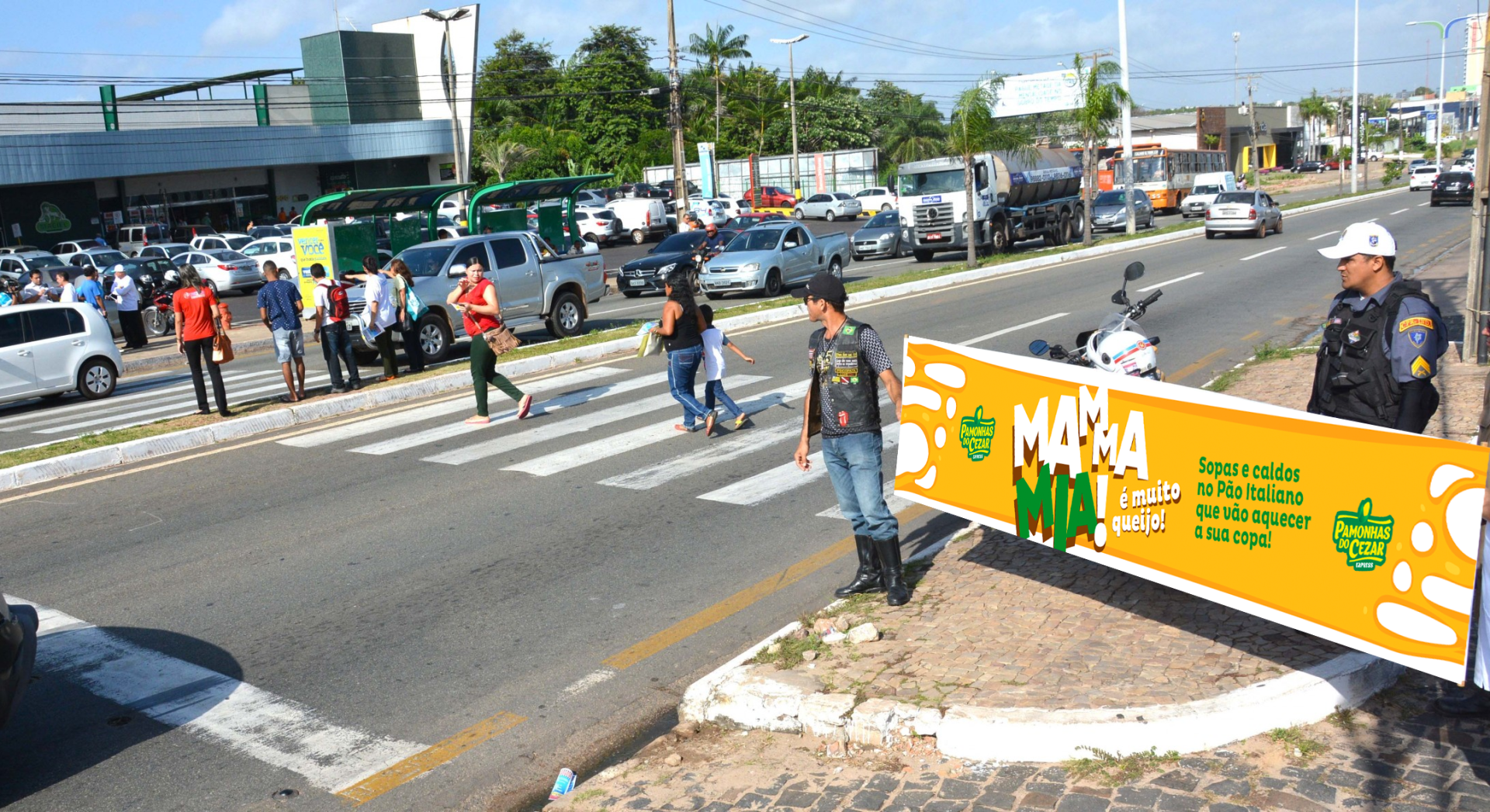 Pessoas atravessando a rua na faixa de pedestres, um policial parado ao lado de uma placa. Carros estacionados na rua.