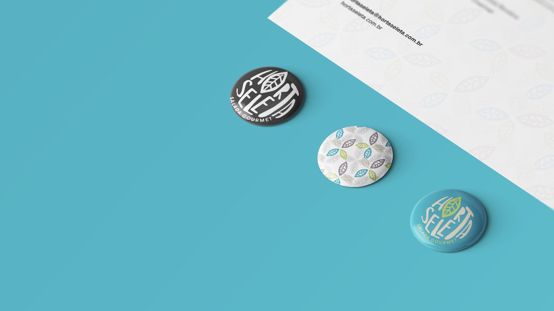 Três ímãs redondos sobre uma superfície azul, ao lado de um papel branco parcialmente visível. Os ímãs têm diferentes designs e cores: preto e branco, estampados e azul-claro.