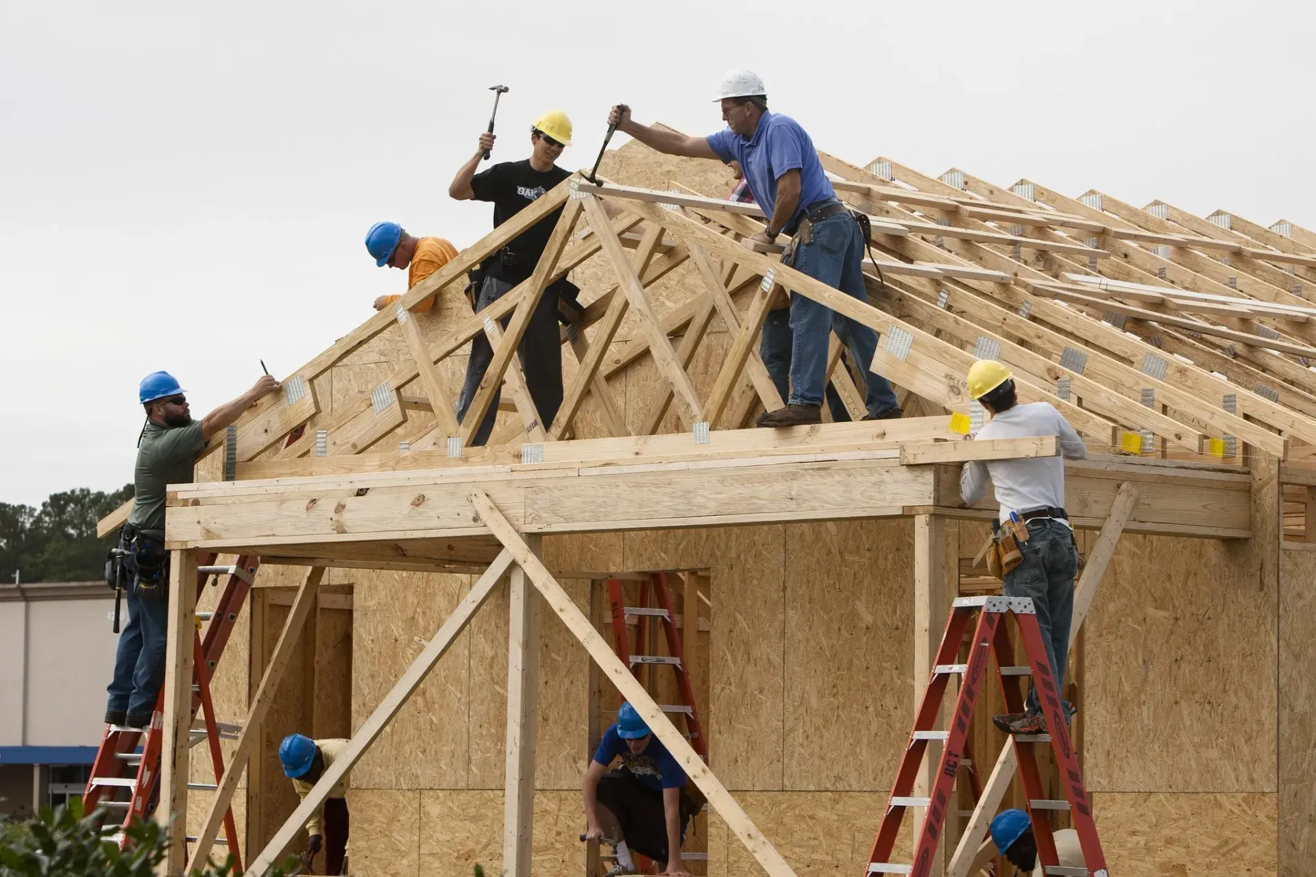 Construction workers build a wooden roof frame on a house, working on ladders and the roof itself.