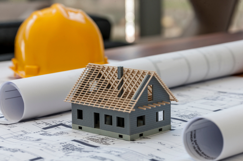 A model house is sitting on top of a blueprint next to a hard hat.