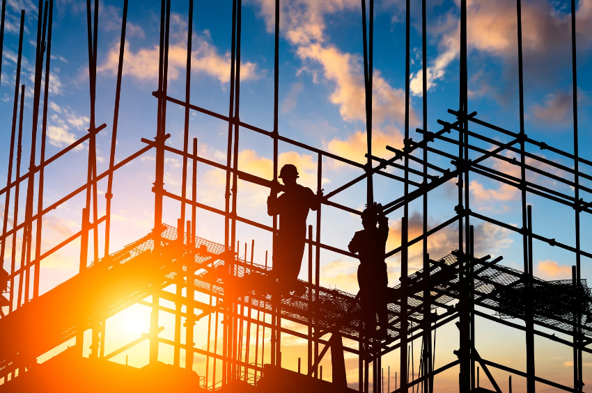 Two construction workers are standing on scaffolding at a construction site at sunset.