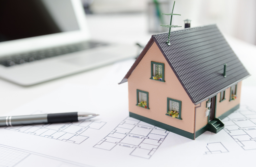 A model house is sitting on top of a blueprint next to a pen and a laptop.