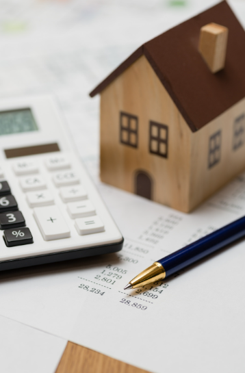 A wooden house sits next to a calculator and a pen