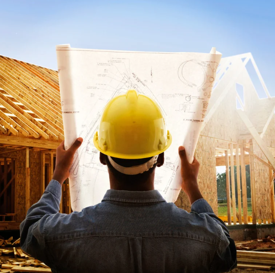 A man wearing a hard hat is holding a blueprint in front of a house under construction