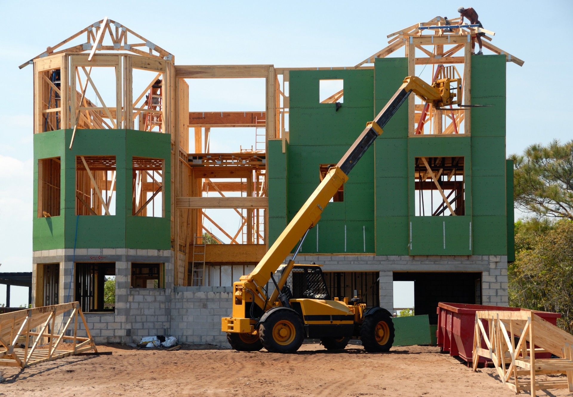 Construction of a two-story house with green siding, wooden frames, and a yellow forklift.
