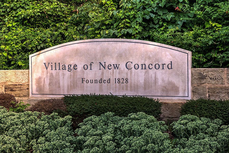 Sign for the Village of New Concord, founded 1828, set in a stone wall and greenery.