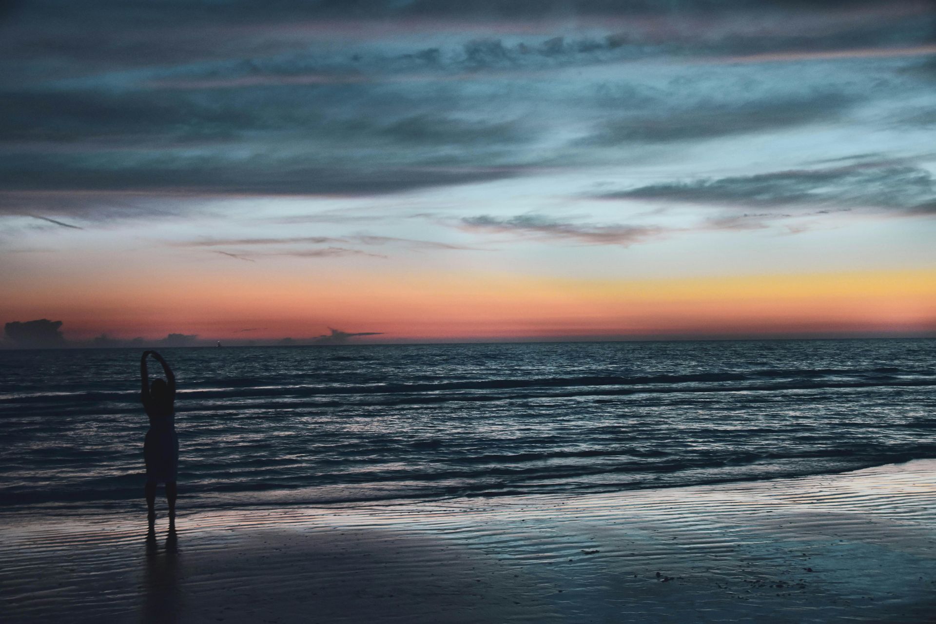 A person is standing on a beach holding a surfboard at sunset.