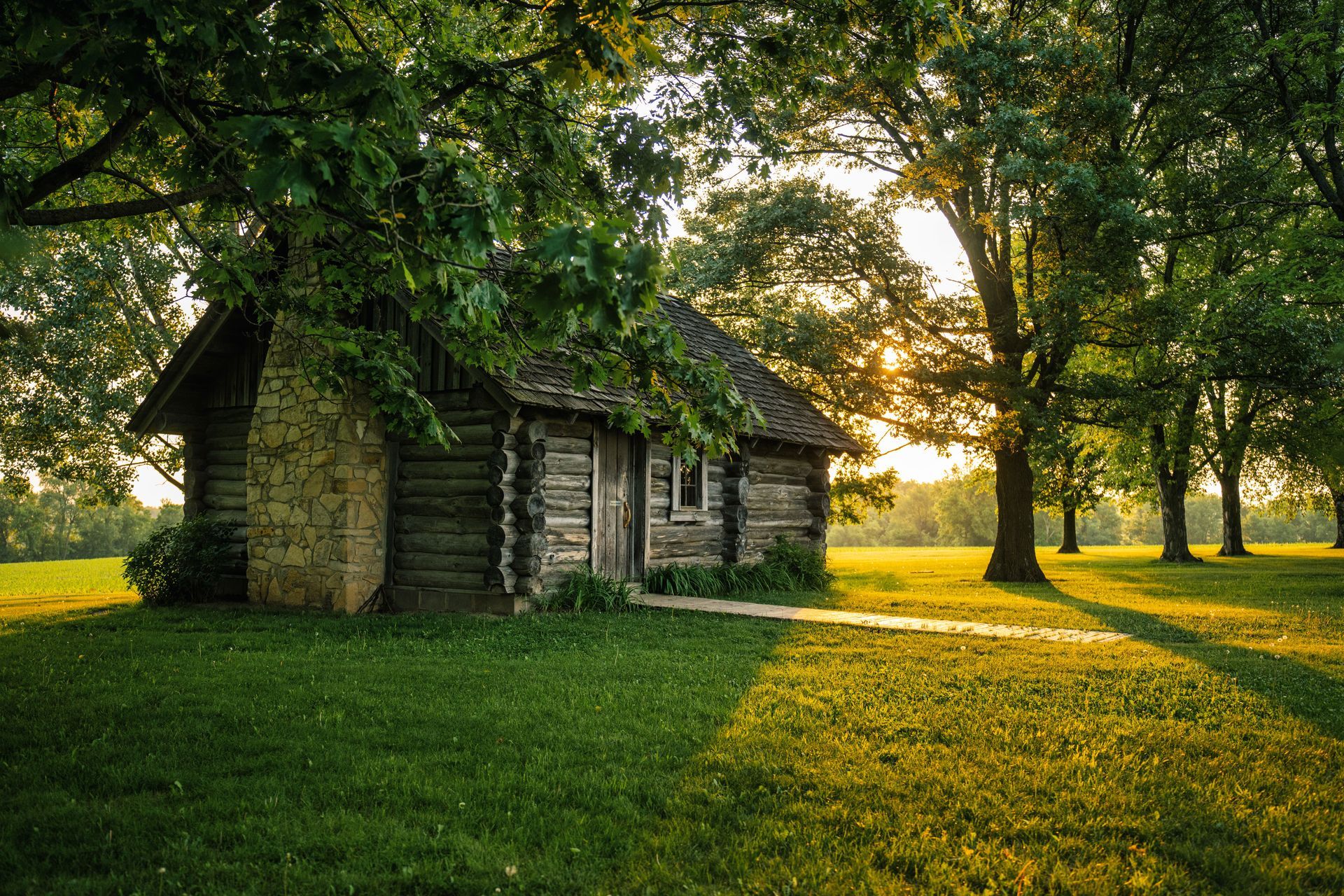 Stone cabin in a sunlit grassy grove, with long golden shadows under leafy trees.