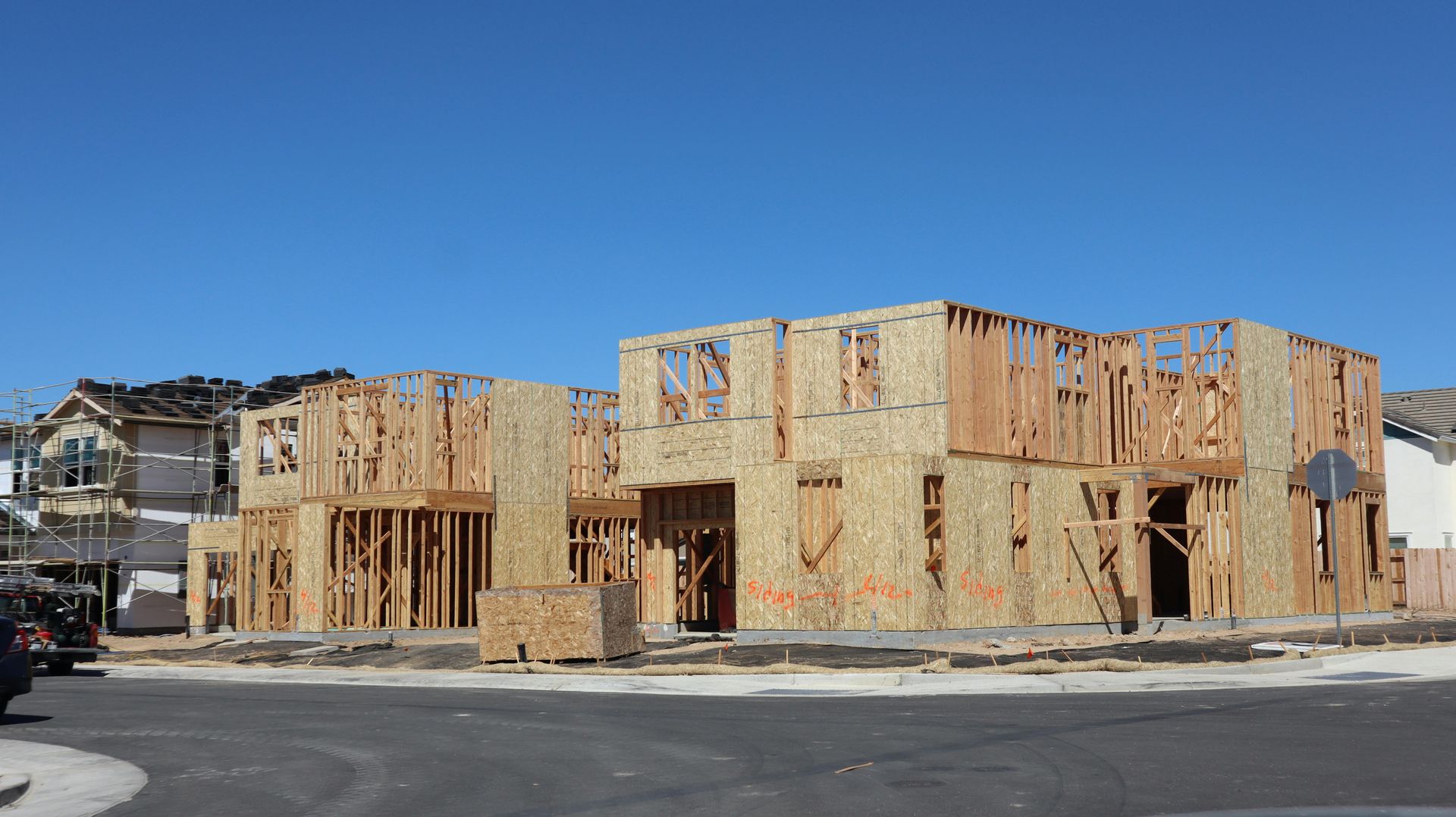 A multi-unit residential building under construction, featuring wooden framing and plywood siding against a clear blue sky.