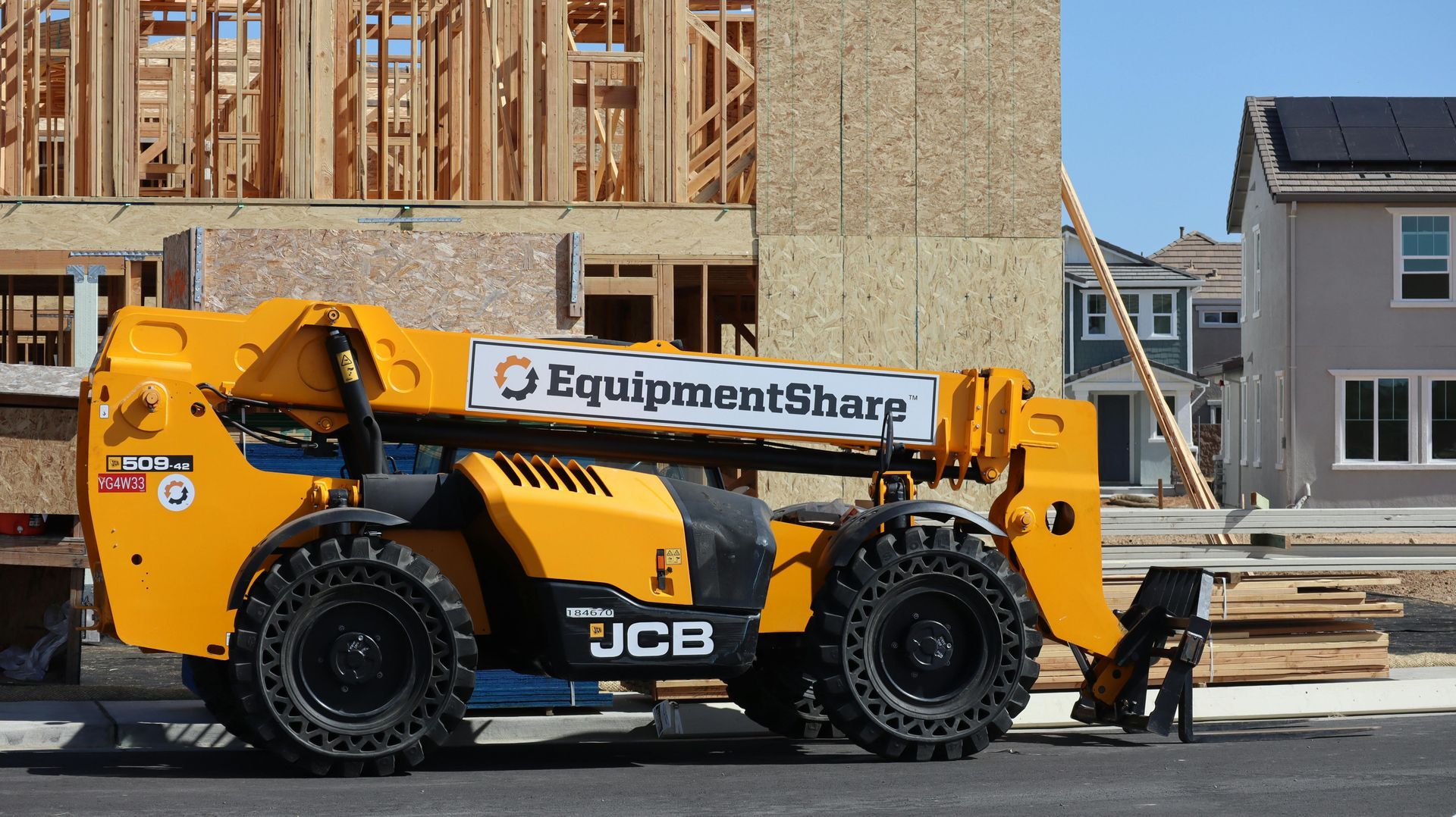 Yellow telehandler at construction site, with wooden structure and houses in the background. EquipmentSharp logo visible.