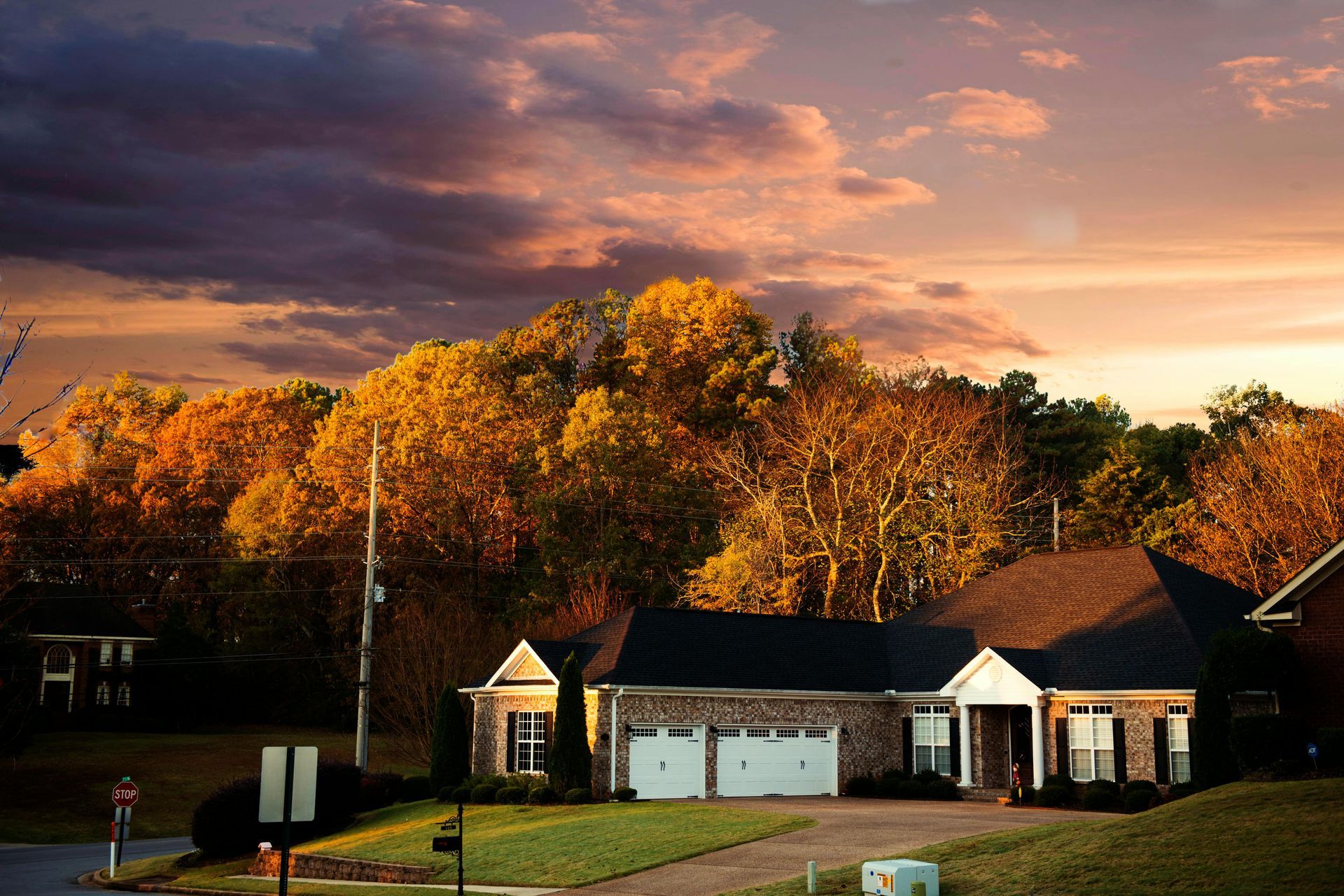 House with a two-car garage at dusk, surrounded by trees with autumn foliage, under a colorful sky.